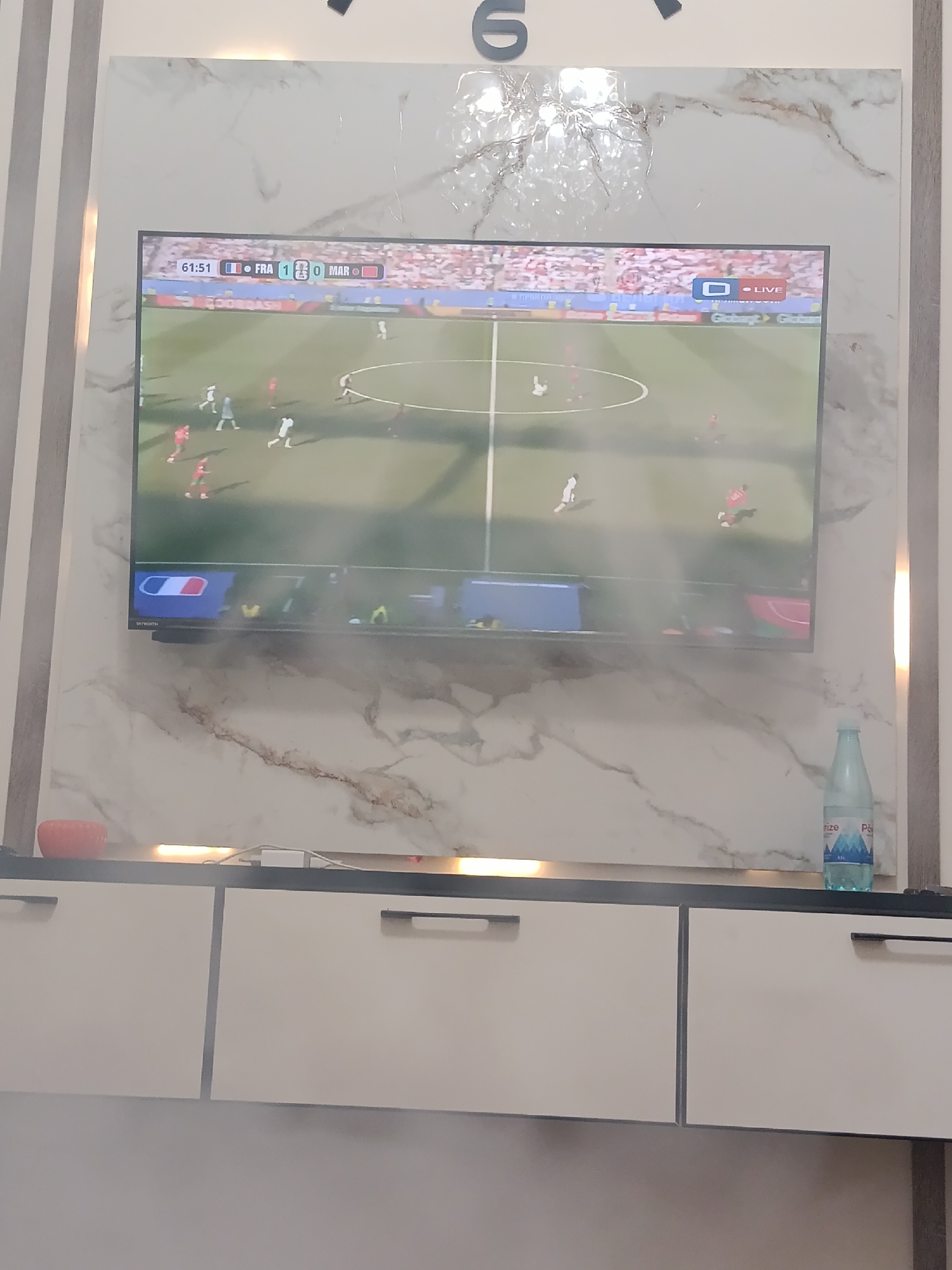 The image shows a television screen displaying a soccer match. The score reads "61:51 FRA 1-0 MAR". The television is mounted on a wall with a marble-like pattern. Below the television are white cabinets. On the surface above the cabinets, there is a small orange bowl on the left and a bottle of water on the right. The lighting appears to be artificial, with a strip light illuminating the surface below the television. There is no specific indication of the location within the city of Ashgabat, Turkmenistan.
