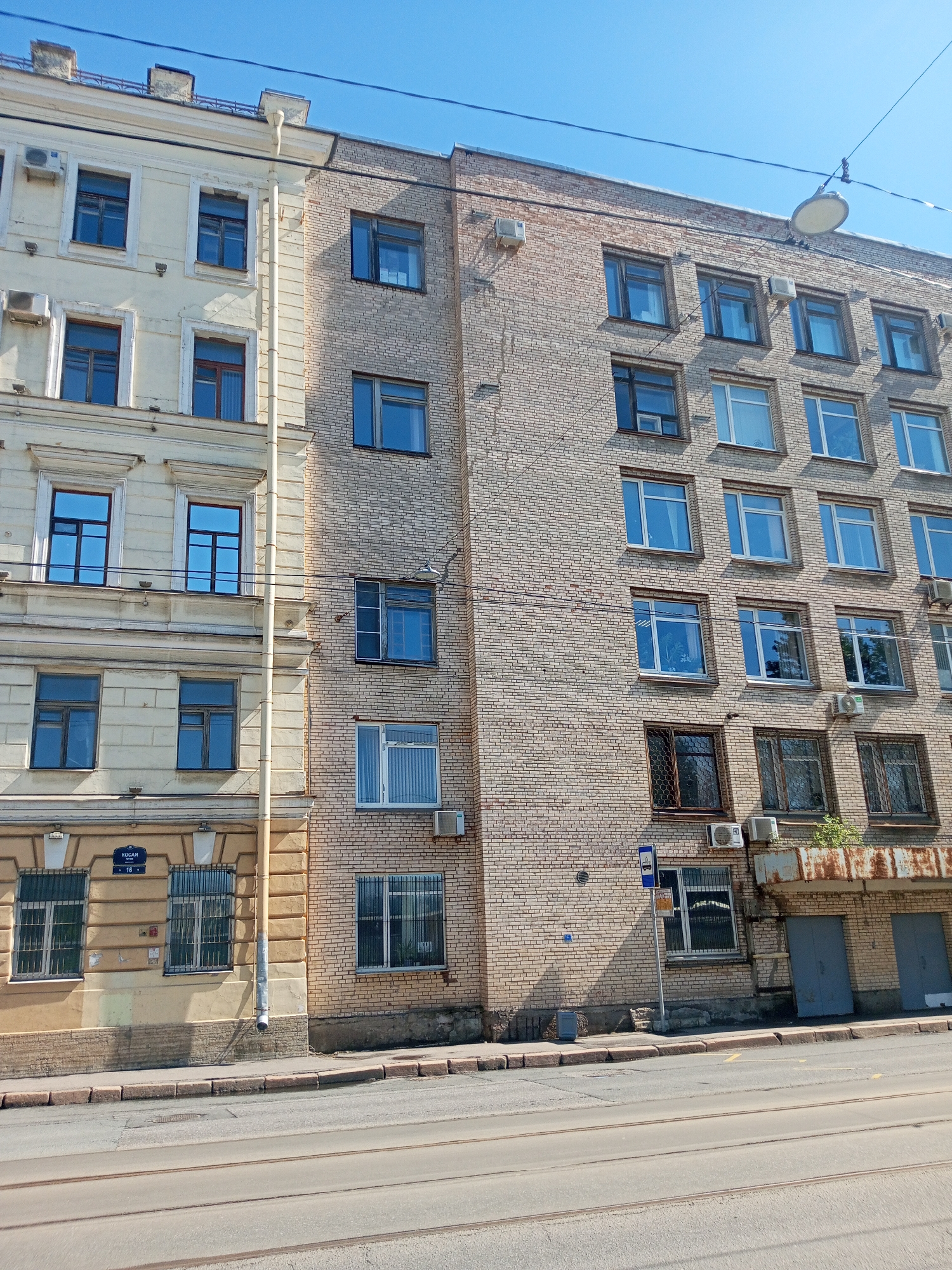 The image depicts two adjacent buildings in Saint Petersburg, Russia. The building on the left is a light yellow color with ornate trim and several windows with bars. The building on the right is made of light tan brick with numerous windows, many with air conditioning units installed below them. There is a street sign on the left building. In front of the buildings is a road with tram tracks. The sky is clear and blue.