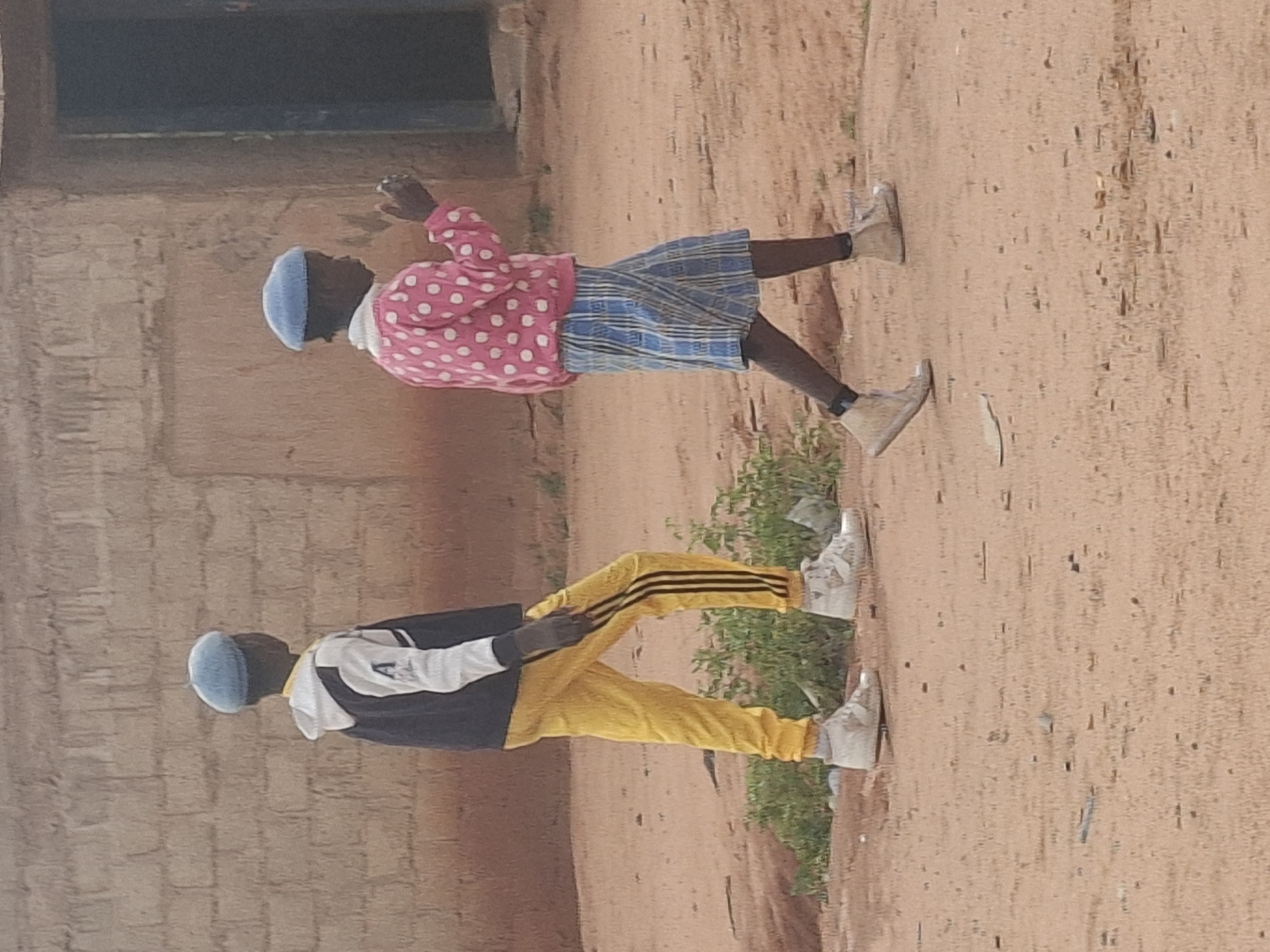 Two young individuals, likely children, are walking outdoors on a sandy surface. The individual on the left wears a dark jacket with white sleeves and the letter 'A' on the chest, paired with bright yellow pants featuring black stripes down the sides. They also wear white sneakers. The individual on the right is dressed in a pink sweater with white polka dots and a blue and white plaid skirt. They are also wearing light-colored sneakers. Both individuals are wearing light blue berets. The background shows a brick building with a dark doorway. The scene appears to be in an outdoor, possibly rural or semi-urban environment. The general impression is a candid, unposed moment of two children walking. This scene is set in Tumfure, Nigeria.
