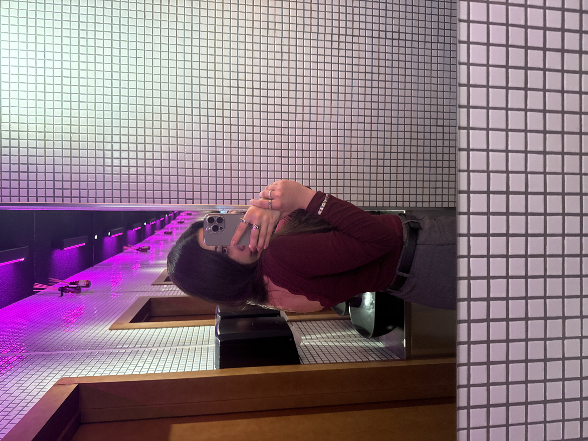 A woman is taking a selfie in a bathroom that has white tiled walls and a long counter. The woman has long, dark hair and is wearing a maroon long-sleeved shirt and grey pants. She is holding her phone in her hands to take the picture. The counter has a built-in sink, and there are reed diffusers on the counter. The lighting in the bathroom is a mix of white and purple. There is a mirror in front of her. The location is likely Yerevan, Armenia.
