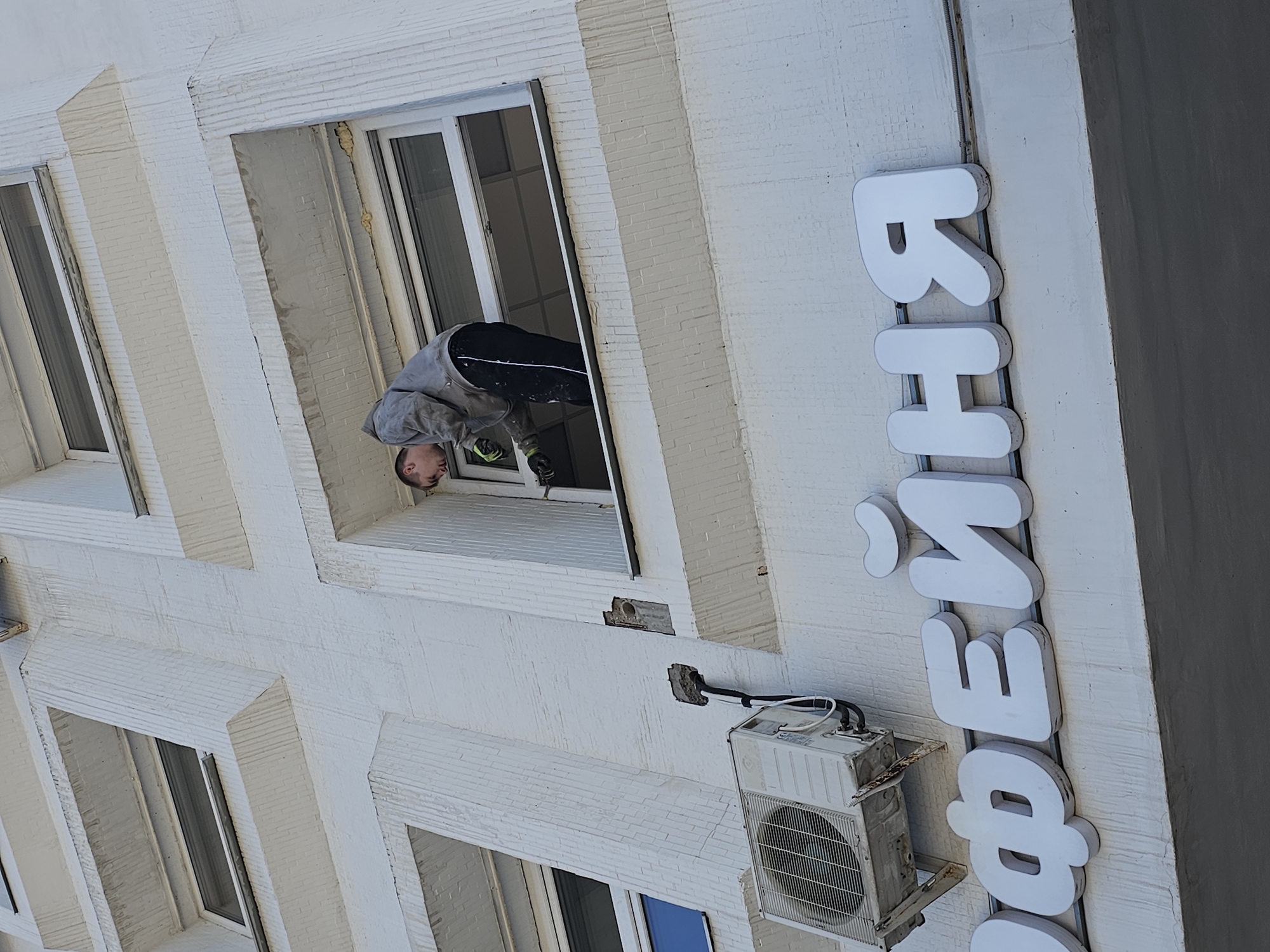 A man is leaning out of a window on a building in Moscow, Russia. The building is white and has several windows. A sign on the side of the building reads "ВЕНИСЕ". An air conditioner is mounted on the building below the window the man is leaning out of. The man is wearing a gray sweatshirt, black pants, and gloves. He appears to be working on something outside the window.