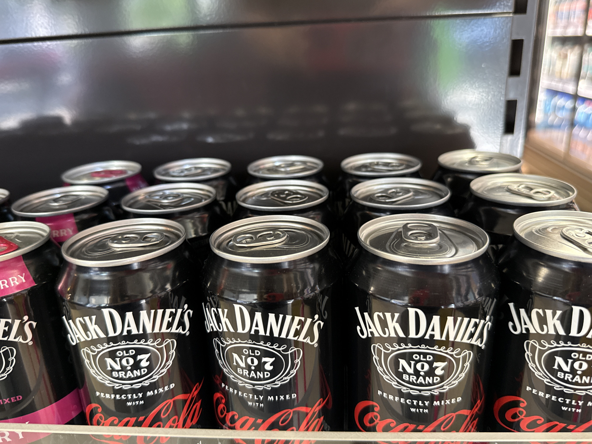 The image shows a close-up, eye-level, medium shot of a shelf stocked with cans of Jack Daniel's Old No. 7 Brand perfectly mixed with Coca-Cola. The cans are primarily black with white and red lettering. The Jack Daniel's logo, a white oval with "OLD NO. 7 BRAND" in the center and decorative swirling lines above and below, is prominent. Below this, "PERFECTLY MIXED WITH" is written, followed by the red Coca-Cola logo. Some cans in the foreground have slightly tilted tops, indicating they are on a shelf. To the left, partially visible, are cans with pink accents and the word "ERRY" visible, suggesting a different flavor of Jack Daniel's mixed drink. The background is a blurred, dark, reflective surface, possibly the interior of a cooler or shelf. Further back on the right, out of focus, are rows of other beverages on shelves, appearing to be in a retail environment. The scene is likely indoors, possibly a convenience store or supermarket. The context of the city of Bad Lauterberg im Harz, Germany, is not directly discernible from the visual information in the image.