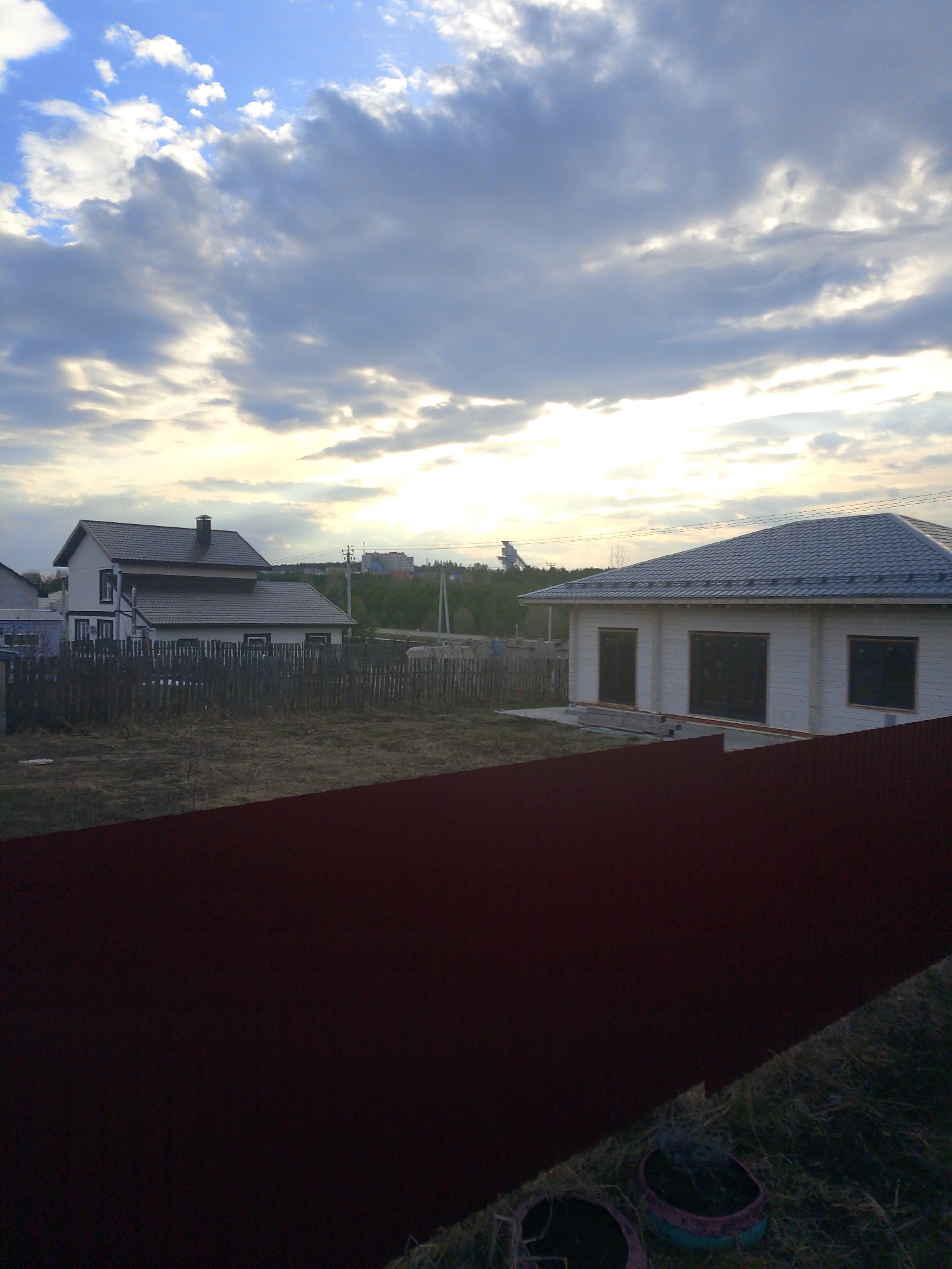 The photograph captures a view in what appears to be the outskirts of the city of Прикамский, Russia. Two houses are visible; the one on the left has a dark roof, a chimney, and a fence surrounding it. The house on the right has a light-colored exterior with dark-framed windows and a grey roof. In the foreground is a red metal fence, partially obscuring the view. Beyond the houses, there's a line of trees and what appears to be urban development in the distance. The sky is cloudy, with light breaking through, suggesting either sunrise or sunset. There are some discarded pots visible near the bottom of the frame. The scene has an unkempt and somewhat desolate feel.