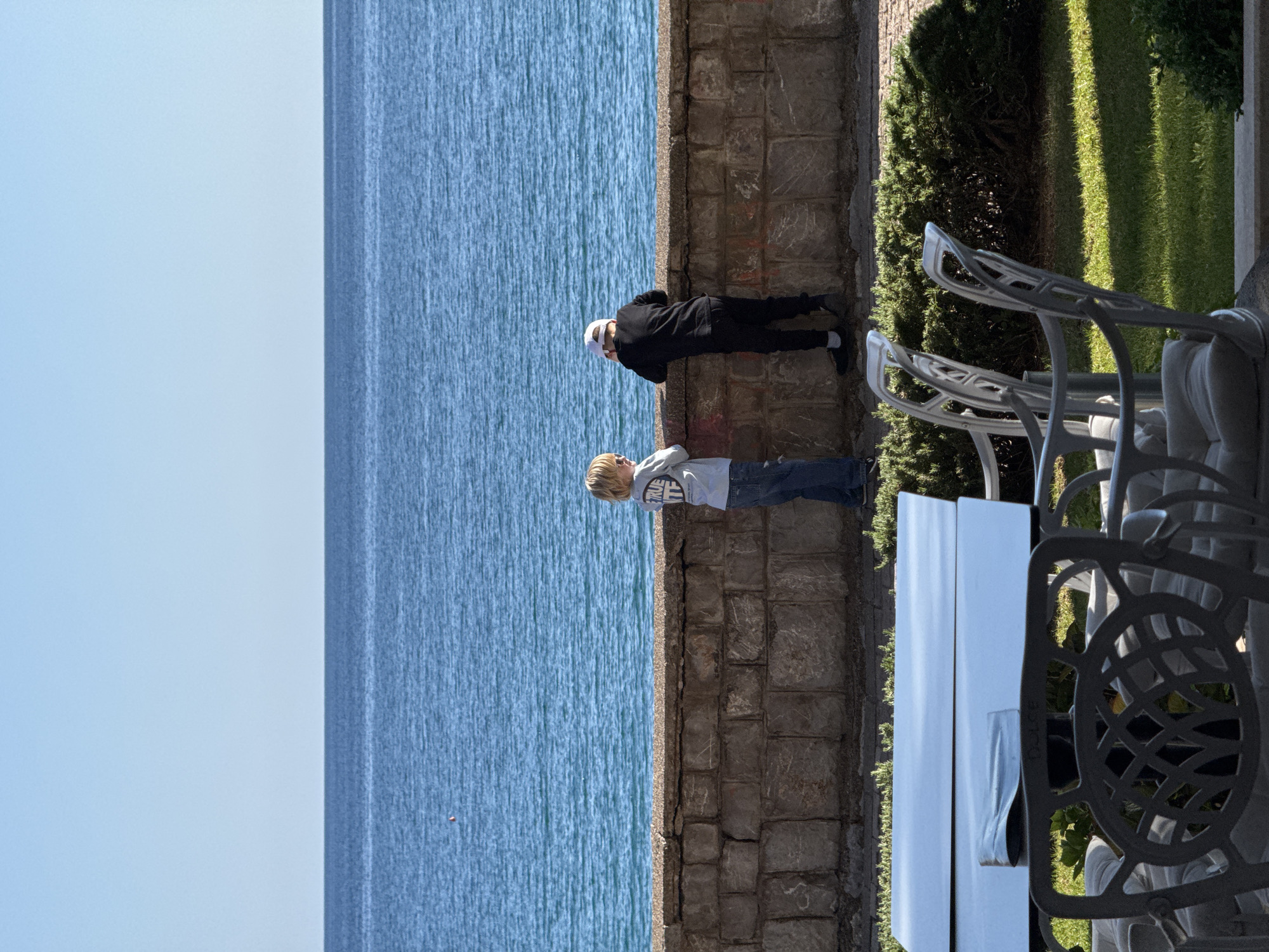 The image shows two figures, presumably children, standing next to a stone wall overlooking a body of water. The water appears to be a large lake or sea, with a clear blue sky above. The wall is constructed from roughly hewn stones, showing signs of age and weathering. Next to the wall, on the right side of the image, is a table with chairs, suggesting a cafe or restaurant setting. Beyond the tables and chairs, a patch of greenery is visible, possibly a garden or park. The location appears to be a coastal city or town with tourist infrastructure.