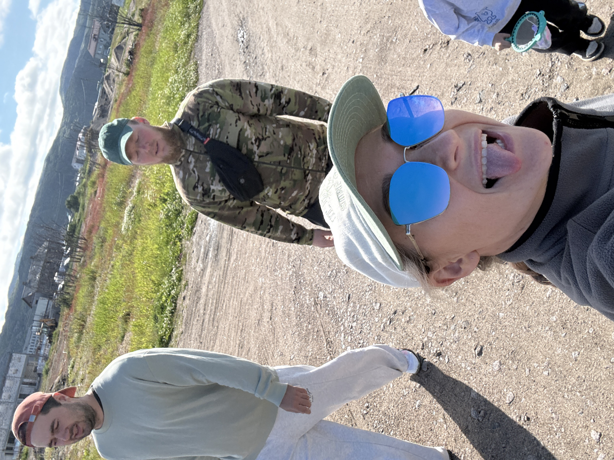 In Teriberka, Russia, on a dirt road, a group of four people is pictured. In the foreground, a woman wearing a gray jacket, a light green baseball cap, and blue mirrored sunglasses is taking a selfie, sticking her tongue out at the camera. To her right and slightly behind, a child in a light-colored sweatshirt and dark pants holds a green butterfly net. In the mid-ground, two men are walking. The man on the left wears a light green crewneck sweatshirt and gray sweatpants, and a reddish-brown baseball cap. The man on the right is wearing a camouflage jacket with a black fanny pack and a dark green baseball cap. The background shows a hilly landscape with some sparse vegetation and unfinished buildings, characteristic of a coastal town under development. The sky is partly cloudy and blue.