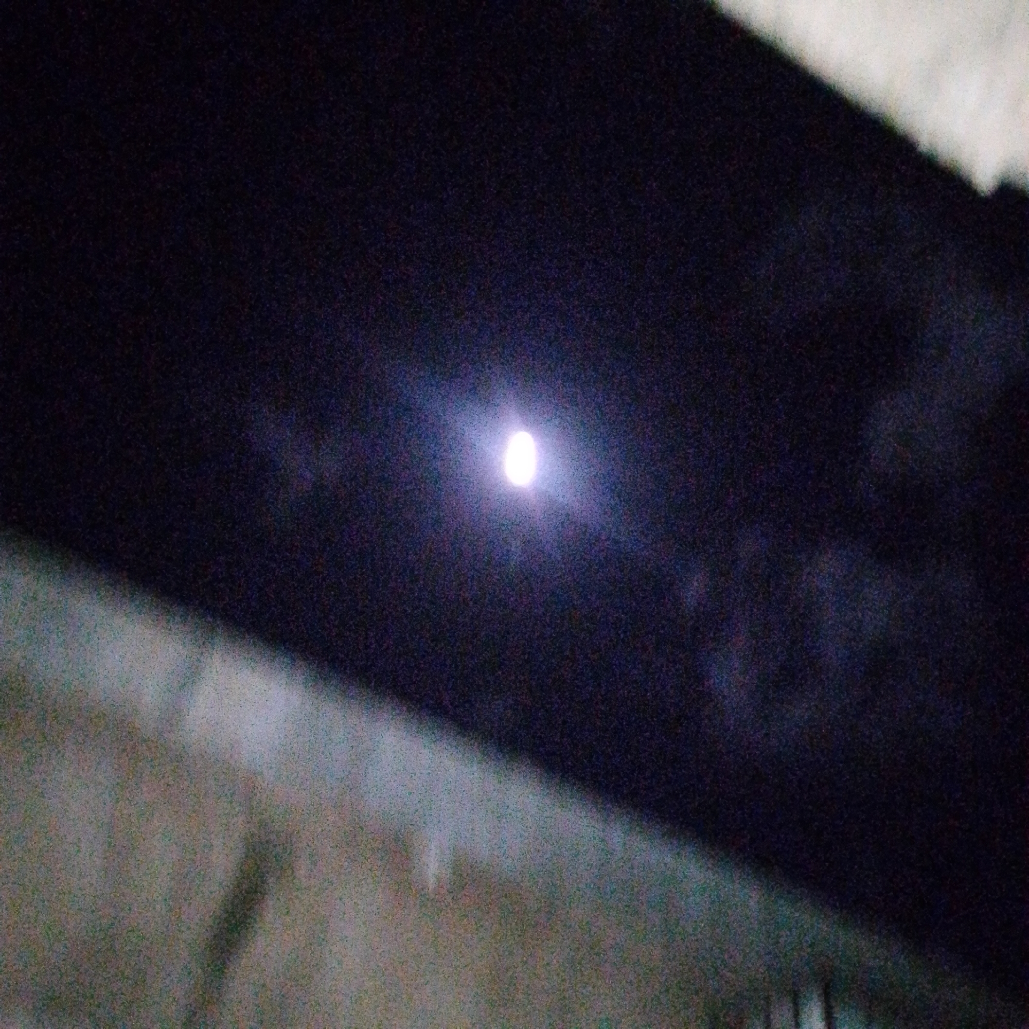 The image, taken at night in Altagracia, Venezuela, shows a small, bright light source centered against a dark, possibly interior, background. The light is surrounded by a hazy, almost diffused glow. In the lower left, a rough, light-colored surface is visible, possibly a wall or the ground. The upper right contains a dark, angular shape, possibly a building or other structure. The image is grainy and lacks detail due to low light conditions.