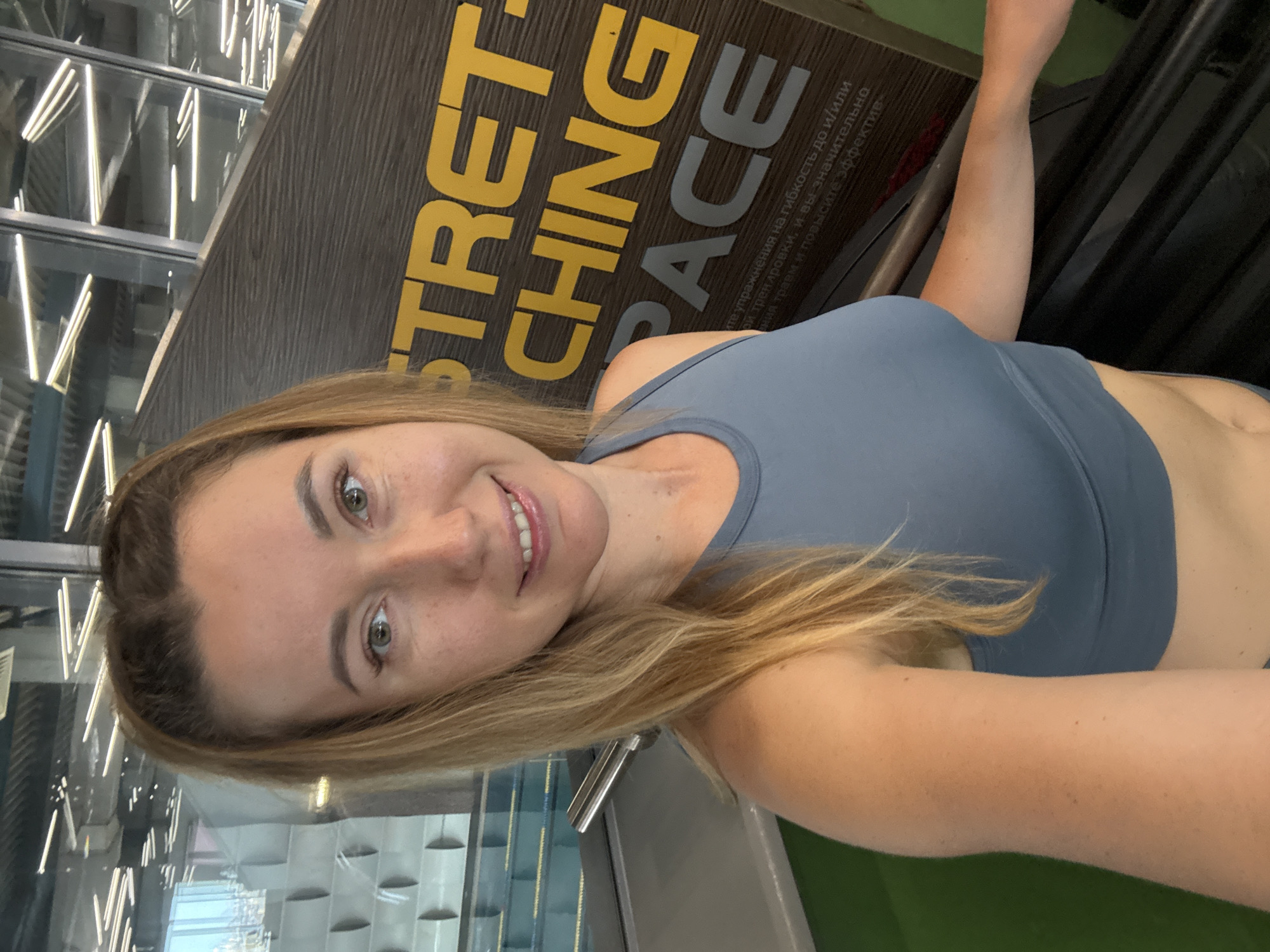 A woman with blonde hair pulled into a ponytail takes a selfie in a gym. She is wearing a blue sports bra. Behind her, a sign reads "STRETCHING SPACE" in yellow and gray lettering. In the background, there is a swimming pool and a modern building interior with fluorescent lighting. The woman is smiling at the camera.