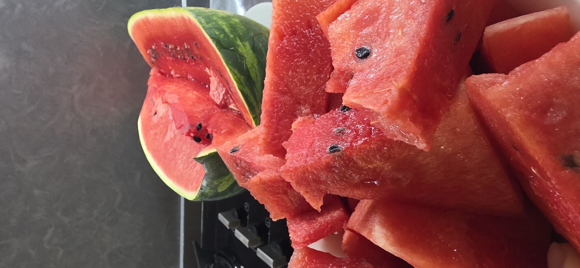 The image displays a close-up view of cut watermelon pieces arranged in a white bowl. A larger, halved watermelon with visible seeds rests on a black surface behind the bowl. The background is a dark, textured wall. The lighting is bright, highlighting the vibrant red of the watermelon flesh. There are no people or any discernible location in Sumgayit, Azerbaijan visible in the image.
