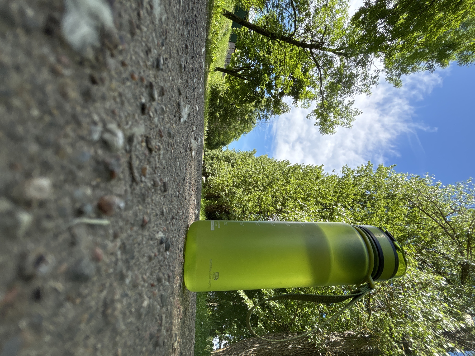 The image shows a bright green water bottle on a gravel path with trees and a blue sky in the background. The perspective is very low, looking up at the bottle and the sky. The trees are lush and green, suggesting a summer day. The sky is a clear blue with some wispy white clouds. The gravel path appears to be well-worn. There are no people visible in the image. The scene appears to be in an outdoor recreational area, possibly a park or a trail. There is no information available to specifically place the location in Minsk, Belarus.