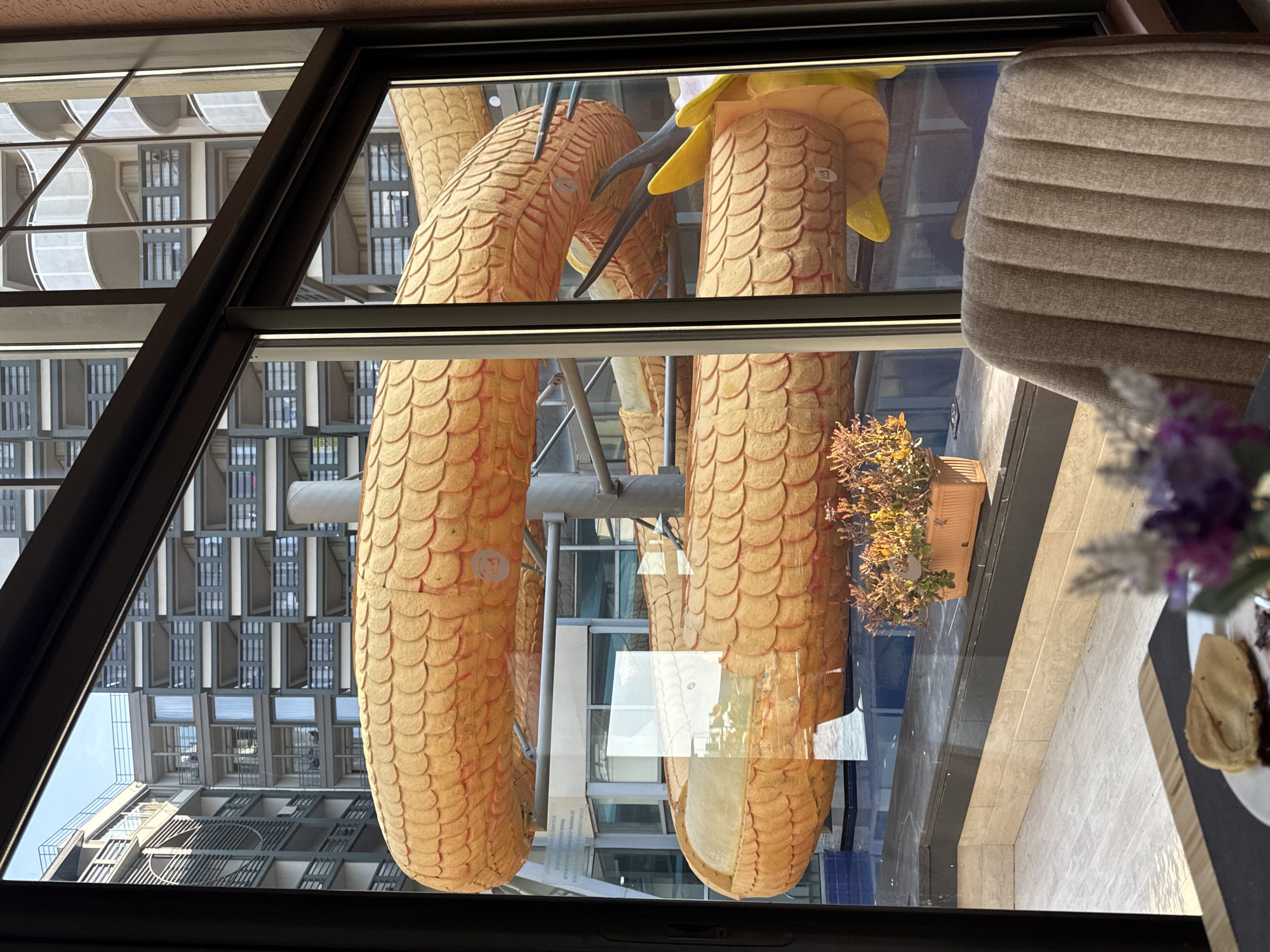 The image was taken in Aksu, Turkey, looking through a window at a large, golden dragon-shaped water slide. The slide is coiled and curves around, with its scales clearly visible. In the background, there are modern buildings with balconies and windows, suggesting a resort or hotel complex. A swimming pool is visible at the base of the slide. Inside the room, a close-up view shows a chair with ribbed upholstery, a table with a plate containing a pancake, and a small vase with purple flowers in the foreground. The lighting suggests it is daytime.
