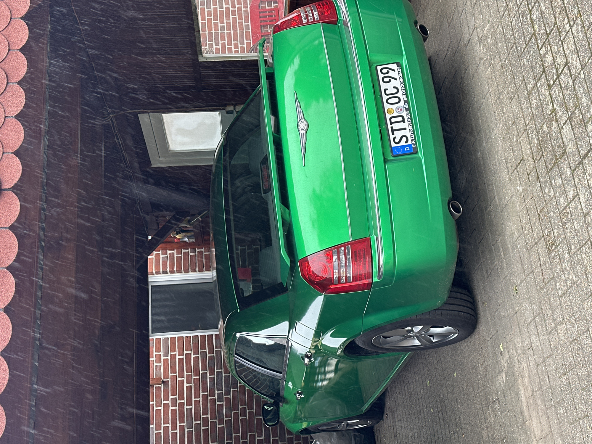 A bright green Chrysler 300C sedan, modified with a rear spoiler and dual exhaust, is parked rear-three-quarter under a wooden carport or lean-to structure. The car bears a German license plate, "STD OC 99," indicating registration in the Stade district of Lower Saxony, Germany, which includes Deinste. Streaks of rain are visibly falling from the scalloped red-tiled roof of the structure. The ground is paved with light grey rectangular concrete pavers. In the background, a building with red brick walls and white-framed windows is visible. To the right, a portion of a red slatted wooden bench can be seen. To the left of the car, a dark bag or object rests on the ground. The scene is illuminated by ambient daylight.