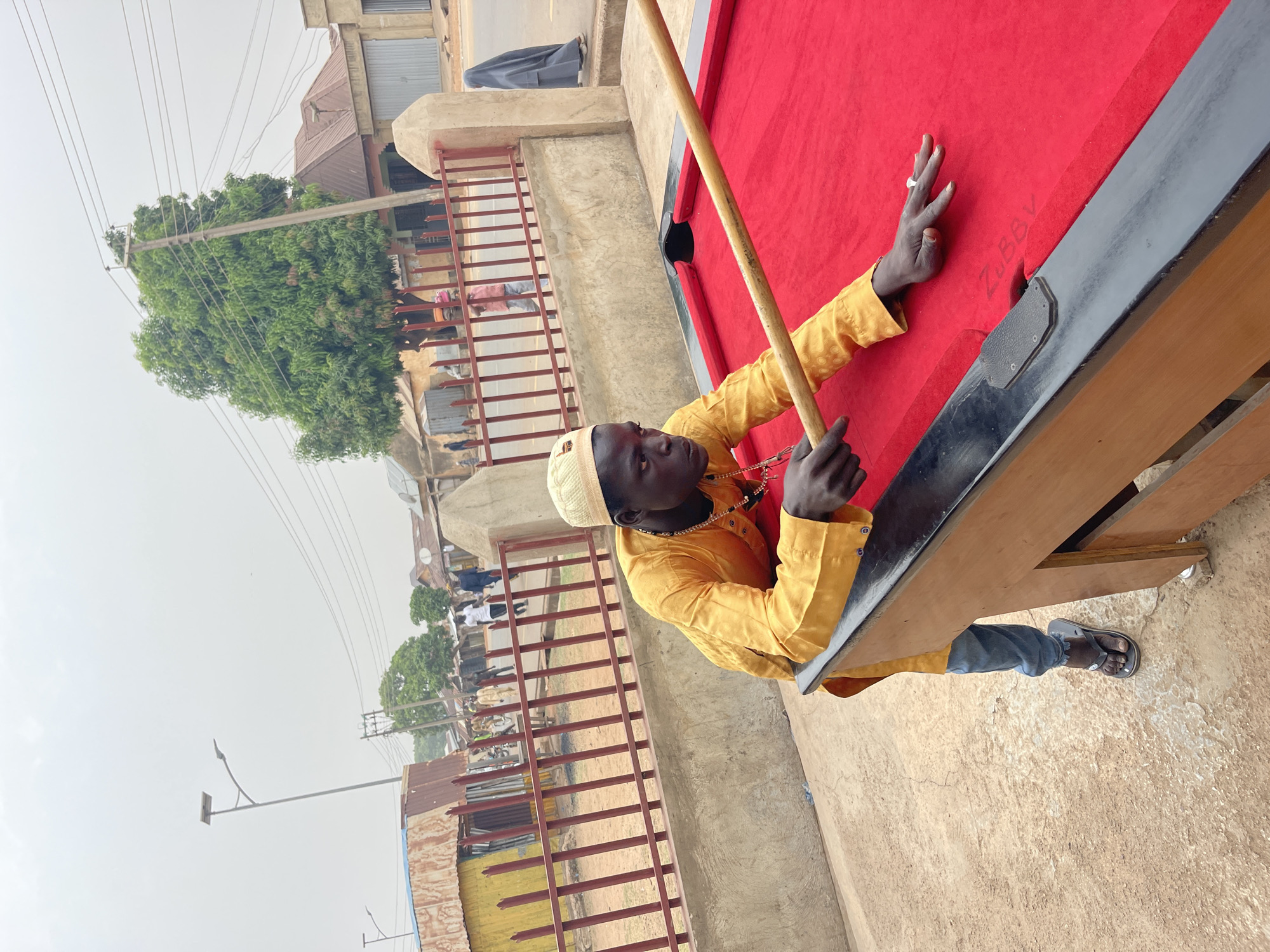 In Unguwar Gabas, Nigeria, a man wearing a yellow shirt and a white cap leans over a red billiard table outdoors. He holds a cue stick in both hands. The billiard table is black-trimmed and located on a weathered surface. Behind the billiard table is a low concrete wall topped with a metal fence. The background shows buildings, trees, and people along the street. A utility pole is also visible. The sky is overcast.