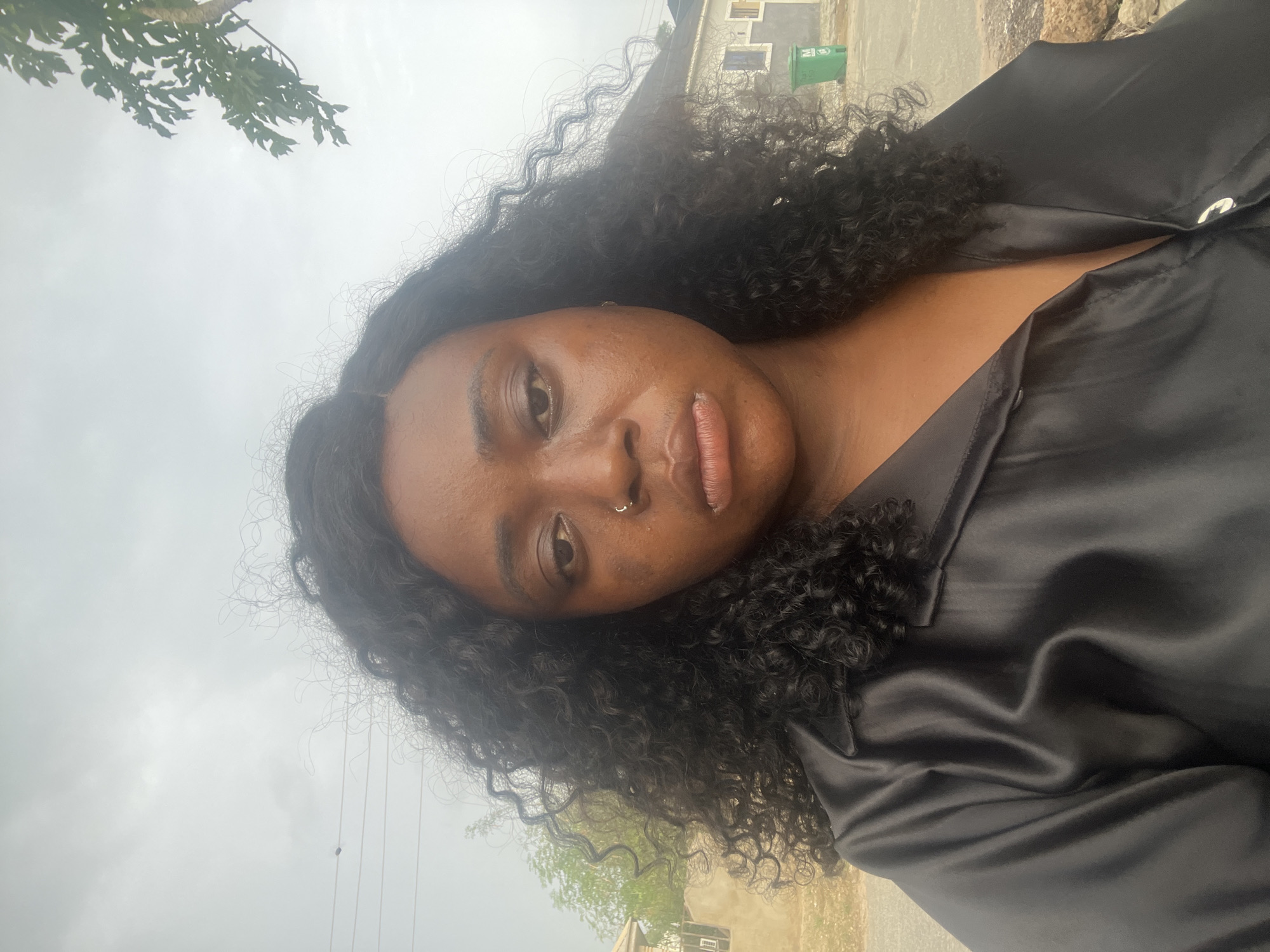 A dark-skinned woman with curly black hair is seen in an outdoor selfie. She has a nose piercing and wears a black satin shirt. The background includes a green bin, a building, trees, and overcast sky. The location is Abuja, Nigeria.
