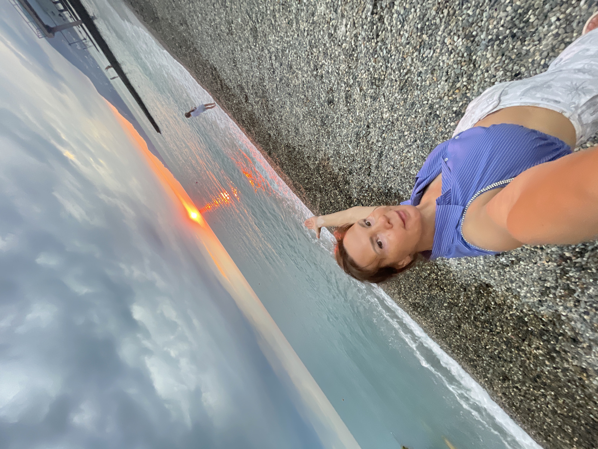 The image shows a selfie taken on a pebble beach in Sochi, Russia, during sunset. The woman in the foreground is wearing a blue striped top and white pants. She is pointing towards the sunset over the Black Sea. In the background, two children are playing in the shallow water. The sky is cloudy with a sliver of orange and red visible where the sun is setting. A pier and mountains are visible in the distance. The overall scene depicts a serene sunset on a beach.