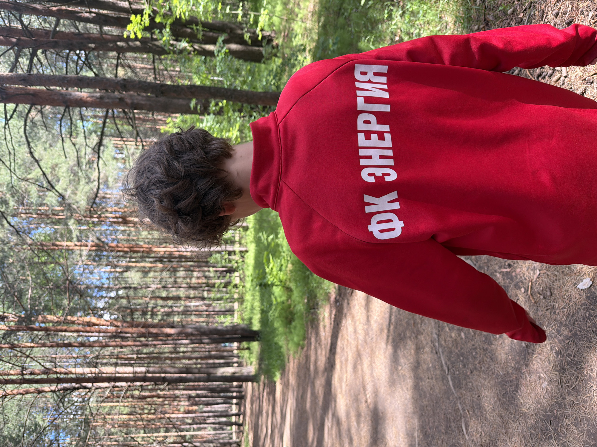 A person wearing a red jacket with white Cyrillic text that reads "ФК ЭНЕРГИЯ" is walking on a path in a forest. The path appears to be dirt or gravel. The location is the city of Chaikovsky, Russia, based on the text on the jacket, which translates to "FC Energia", a football club from that city. The forest is dense with tall, thin trees. The person is facing away from the camera.