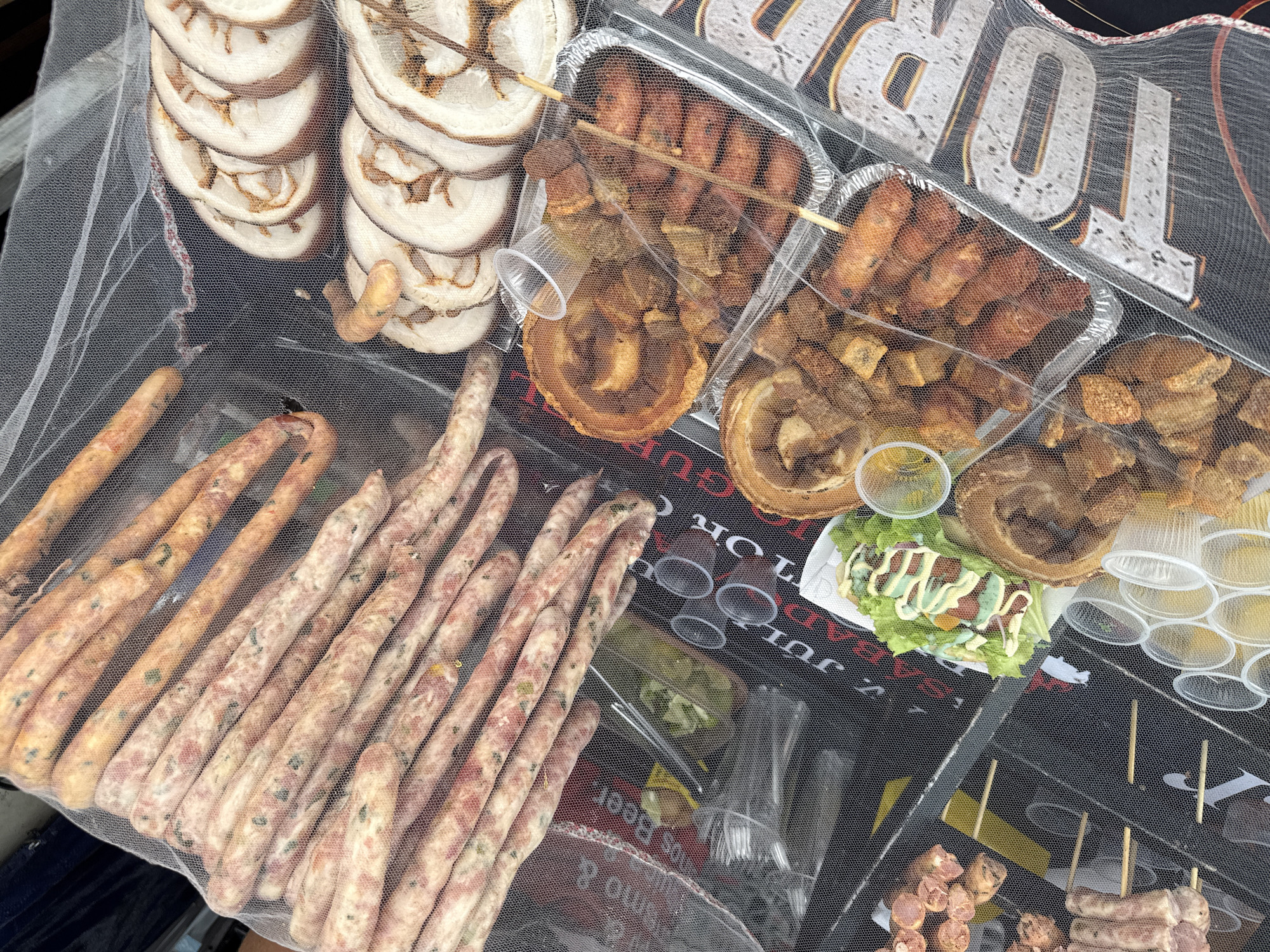 This image depicts an outdoor food stall displaying various meats, likely in a street market setting in Nilópolis, Brazil. A fine white mesh netting covers the food, suggesting protection from insects or dust.
In the upper portion of the image, a large pile of raw, uncooked sausages is visible, densely packed and arranged diagonally. The sausages appear to be about a foot long each and are a pale pinkish-white color with visible flecks of herbs or spices. To the right of the sausages, several slices of cooked pork, possibly porchetta or a similar roast, are arranged in rows. These slices are round with a distinct layered texture of meat and fat, and some appear to have a crispy rind.
Below these items, several foil trays are filled with cooked food. One tray contains what appear to be fried pork rinds or chicharrones, cut into irregular, bite-sized pieces. Another tray holds skewers of grilled meat, possibly sausages or small pieces of pork, with some pieces appearing charred. Adjacent to this, another tray contains more fried pork products. A small white plate holds a salad of lettuce, tomatoes, and a drizzle of dressing, accompanied by a small plastic container of sauce.
Numerous small, clear plastic cups are stacked in various locations, possibly for dips or condiments. The background is dark, with some signage visible through the netting. The overall impression is that of a food vendor preparing or displaying products for sale in a bustling environment.