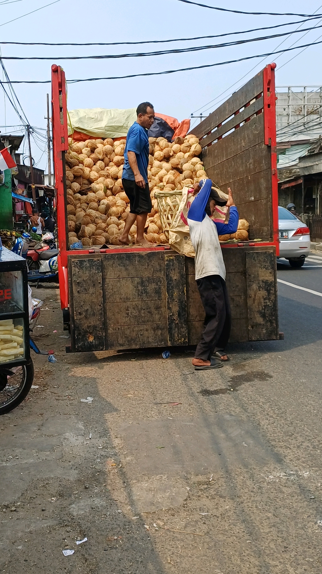 In Citeureup, Indonesia, a truck is parked by the side of the road, its bed filled with a large quantity of coconuts. Two individuals are engaged in handling the coconuts. One man, wearing a blue t-shirt and black shorts, stands in the truck bed, looking to his left. Another man, wearing a white shirt with blue sleeves and black pants, is bending down in front of the truck, his arms raised as if supporting a load or adjusting something. The truck's red wooden sides are prominent, and the coconuts fill the majority of the truck bed, some contained within woven baskets. The surrounding environment appears to be a roadside market area, with stalls, a motorcycle, and a parked car visible in the background. The ground is paved with asphalt and dirt, with scattered debris. The sky is clear and bright, indicating daytime.