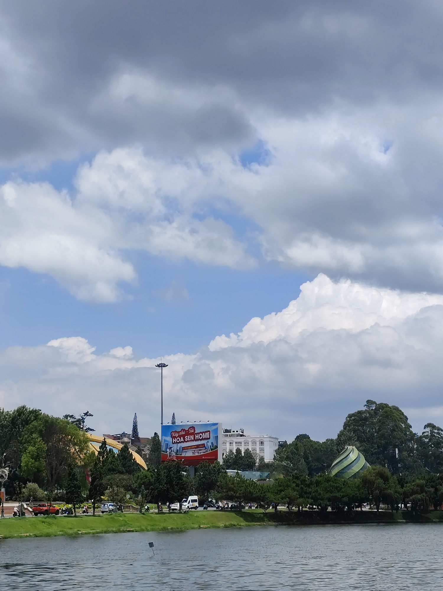 A scenic view of Xuan Huong Lake in Da Lat, Vietnam, under a partially cloudy sky. The lake's calm waters reflect the surrounding landscape. In the background, the distinctive architecture of Da Lat is visible amidst lush trees, including a large billboard that reads HOA SEN HOME. A tall lamppost stands on the shoreline. A road runs along the lake with cars and scooters travelling along it. The green and white building on the right side of the frame is the Dalat Palace Hotel. A spiral-striped geodesic structure is visible on the right side of the frame as well.
