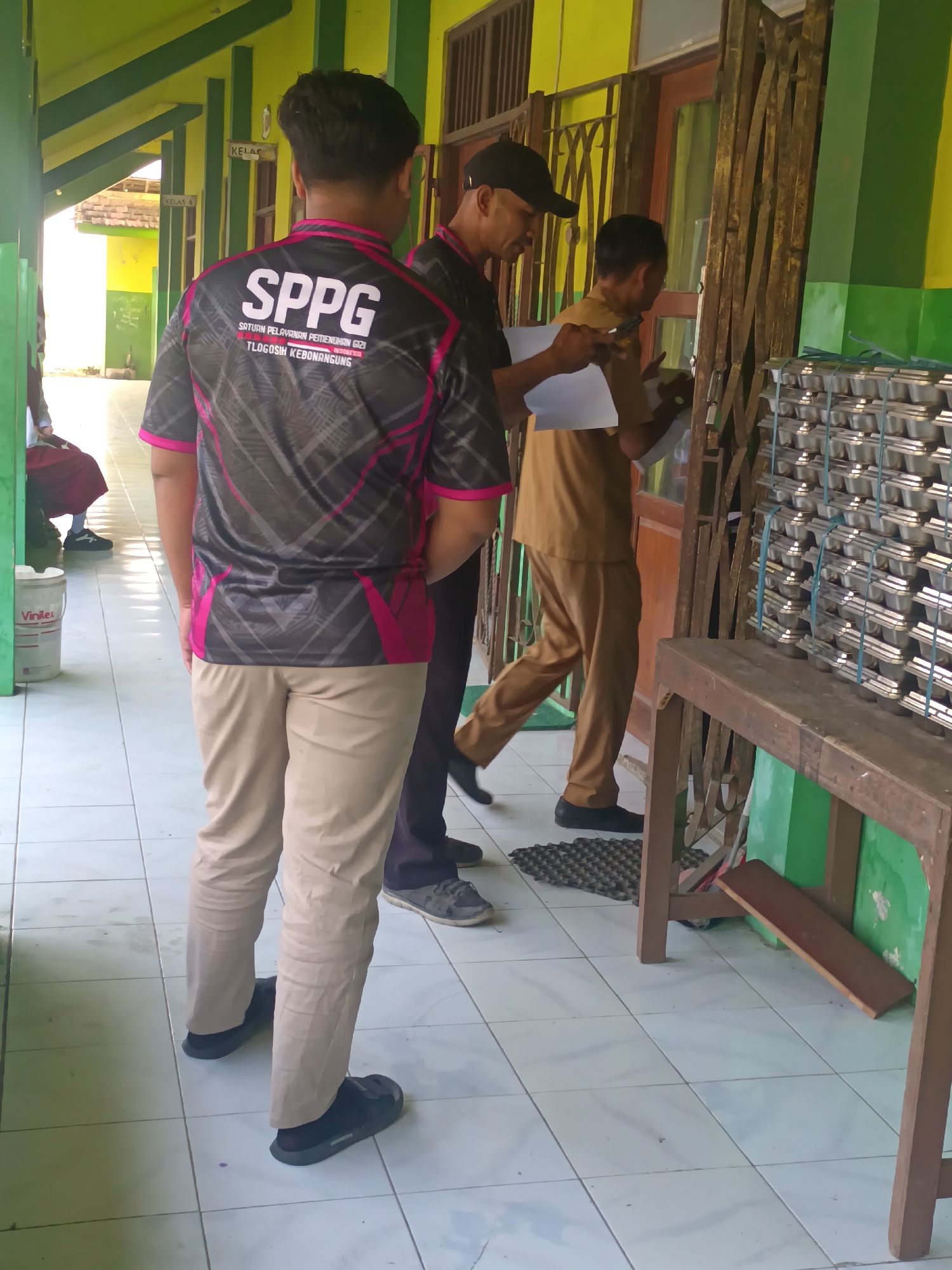 In Sarimulyo, Indonesia, an outdoor hallway with green and yellow walls is depicted. The floor is tiled with white squares. In the foreground, a person wearing a black and pink shirt with the text "SPPG" visible, and khaki pants, stands facing away from the camera. To their right, another person in a brown uniform is seen from the side, holding a piece of paper and interacting with someone partially visible in the background. A third person, wearing a black cap and brown uniform, is also visible, holding a piece of paper. In the background, stacks of what appear to be metal trays or containers are organized on a wooden structure. A metal accordion gate is positioned near the entrance to a building. A bucket with the "Vinilex" logo is on the left side of the image. The scene appears to be set in a school or institutional environment.