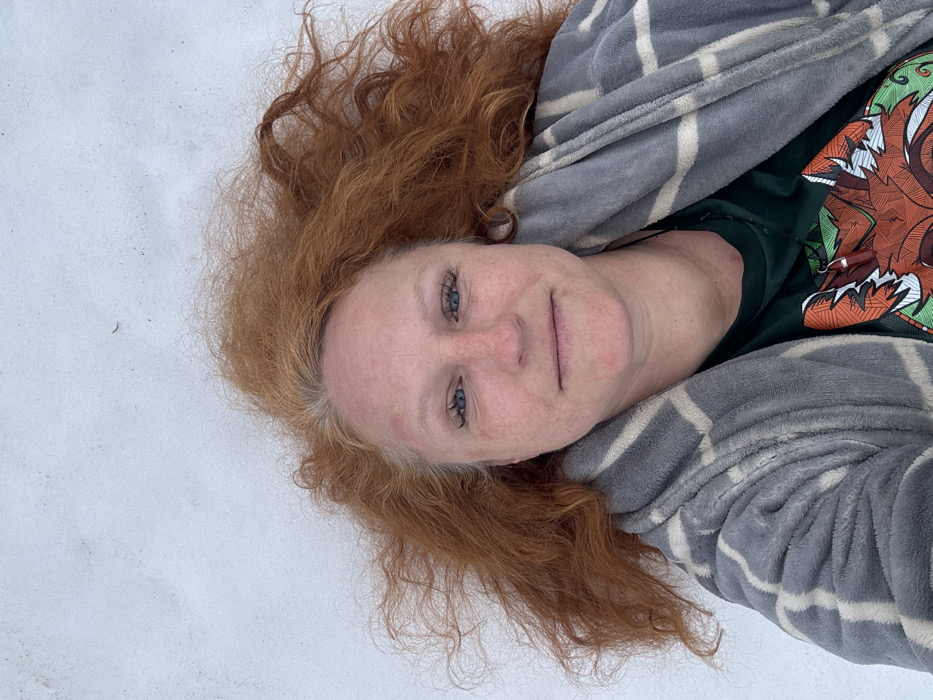 An adult female is depicted from an overhead perspective, lying supine on a white, textured surface consistent with snow. She has long, wavy reddish-orange hair, with visible grey roots, fanned out around her head. Her complexion is fair, with faint areas of erythema on her cheeks and forehead. She possesses blue irises and dark eyelashes and displays a neutral to faintly smiling expression. She is wearing a grey plush robe with a light beige grid pattern, partially open to reveal a dark green t-shirt underneath. The t-shirt features a stylized, multi-colored graphic of a fox's head. A dark string or necklace is visible around her neck. The white background contains a few small dark particulates. The scene is located in the city of Chaikovsky, Russia.
