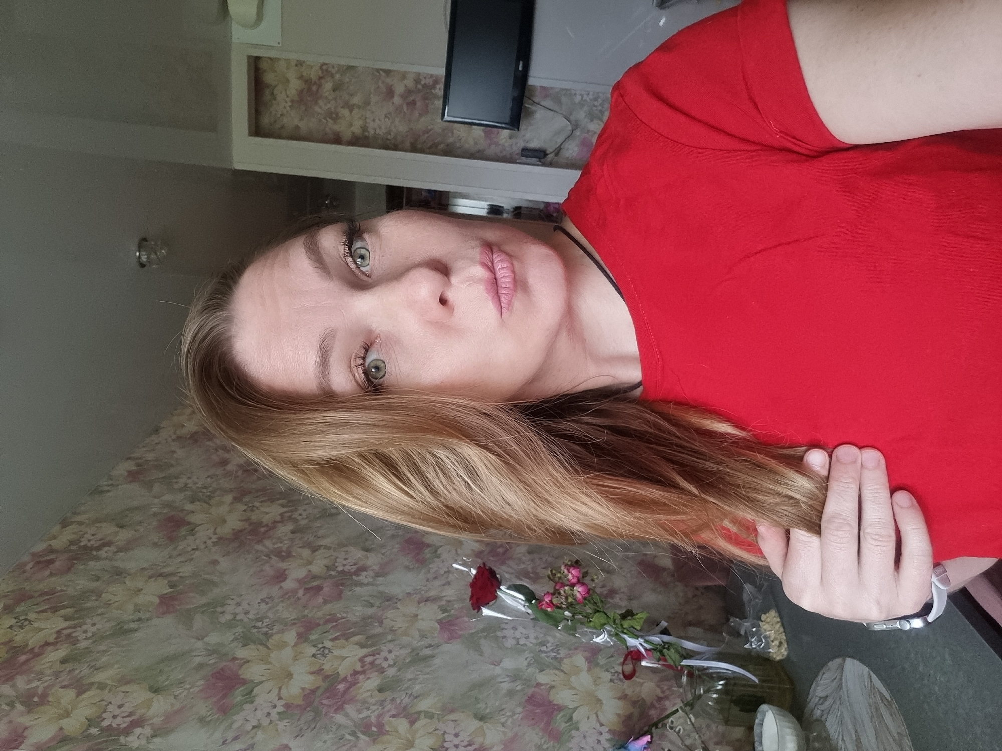The image displays a fair-skinned woman with long, wavy, light brown and blonde hair, and striking light green or hazel eyes. She is wearing a simple red t-shirt and looks directly towards the viewer with a neutral, slightly pensive or pouting expression, her lips lightly tinted pink. Her right hand, with natural nails, gently touches her hair near her shoulder, resting against her red top.
In the lower portion of the frame, to the right of the woman, a small floral arrangement is visible. It consists of a single red rose and several smaller pink or purple flowers, wrapped in clear plastic and tied with a white ribbon. Nearby, parts of a clear glass vase containing dried botanicals, a small white cylindrical object, and a carved light-colored wooden item can be seen.
The background reveals an indoor setting. The wall behind the woman features a classic floral wallpaper pattern with subdued pink, yellow, and green tones. Further back, a flat-screen television is mounted on a plain light-colored wall, situated above a narrow shelf or cabinet. A multi-bulb ceiling light fixture is partially visible near the top left. The overall lighting suggests a well-lit interior during the daytime. The photograph appears to be a posed selfie, capturing a moment within a home in Chaikovsky, Russia.