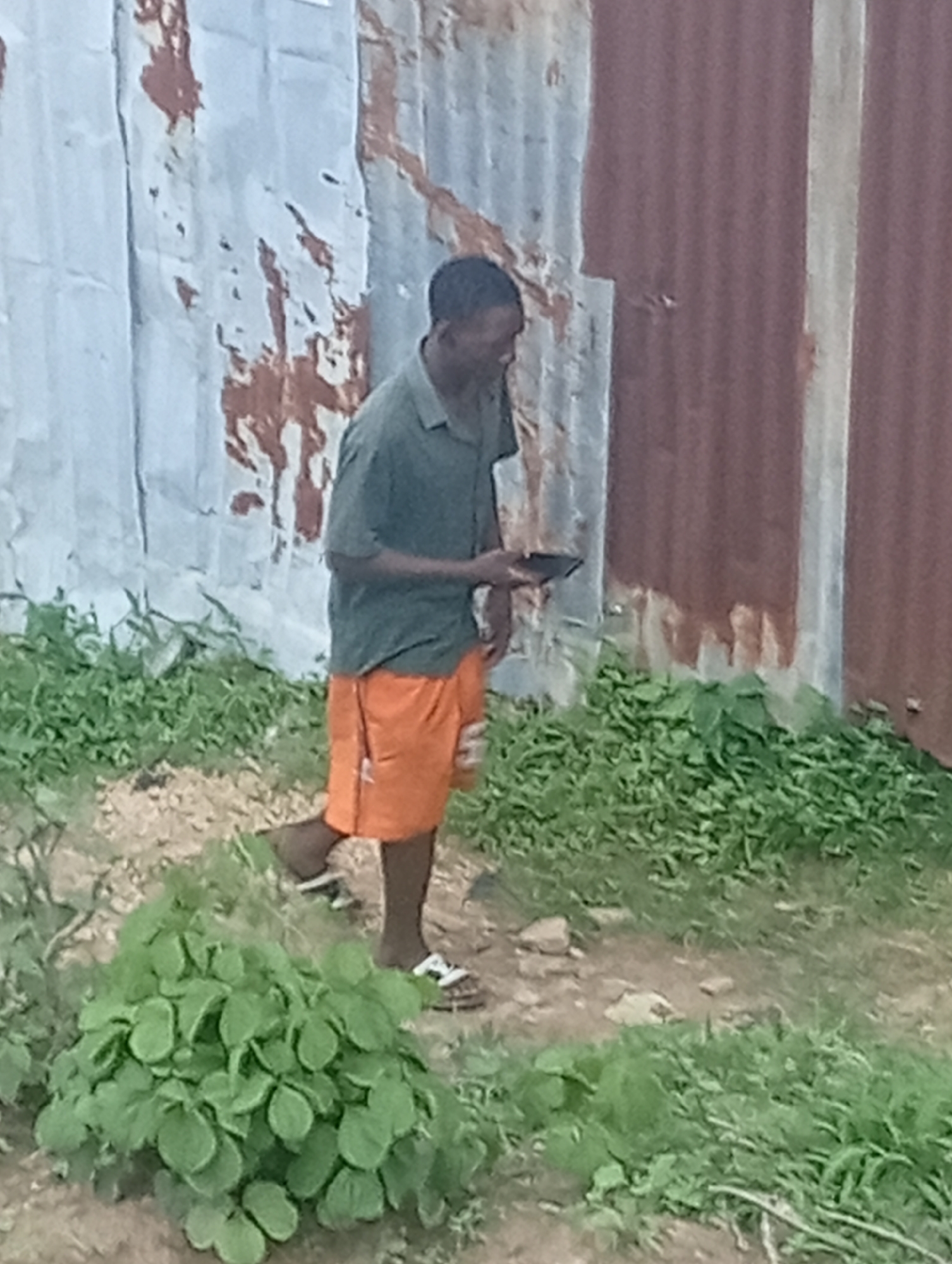 In Jos, Nigeria, a person stands outdoors next to a corrugated metal structure. The individual is wearing a dark green short-sleeved shirt and bright orange shorts. They are holding a dark rectangular object, possibly a phone or tablet, in their right hand, examining it. The person is wearing flip-flops. The ground is dirt with scattered rocks and patches of green foliage. The corrugated structure appears weathered, with rust and paint peeling. The overall scene suggests an informal outdoor setting.