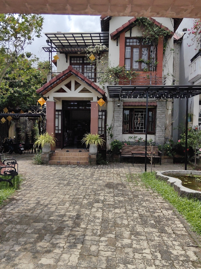 A two-story building, located in Phú Xuân Ward, Vietnam, is viewed from a paved courtyard under an overcast sky. The structure features white-painted walls with reddish-brown tiled gables and roof elements. The central entrance is accessed by three reddish-brown steps, flanked by two gray planters containing vibrant light-green foliage. The doorway is dark and open. Two square, yellow lanterns hang from the triangular-shaped tiled eaves above the entrance.
To the immediate right of the entrance, the ground floor section exhibits a stone-patterned facade with a dark-framed window. A plain wooden bench is positioned against the wall beneath a black metal pergola, from which multiple small white lights are suspended. A security camera is mounted on the wall above this window. The upper floor displays multiple dark-framed windows, with significant green and white-flowering plant growth cascading from a small balcony railing and around the window frames.
The foreground is dominated by a path paved with light-colored, irregularly shaped interlocking concrete pavers, showing areas of discoloration and wear. A narrow strip of green grass borders the paved path on the right, alongside an irregularly shaped, stone-edged pond containing murky water and some surface vegetation. To the far left of the paved area, a dark wooden bench with black metal scrollwork arms is partially visible on the grass. Further left, behind a weathered wall, two dark motorcycles are parked amidst dense green foliage. Additional square yellow lanterns and small white lights are strung among the trees and overhead structures on the left side of the scene. No individuals are present in the frame.
