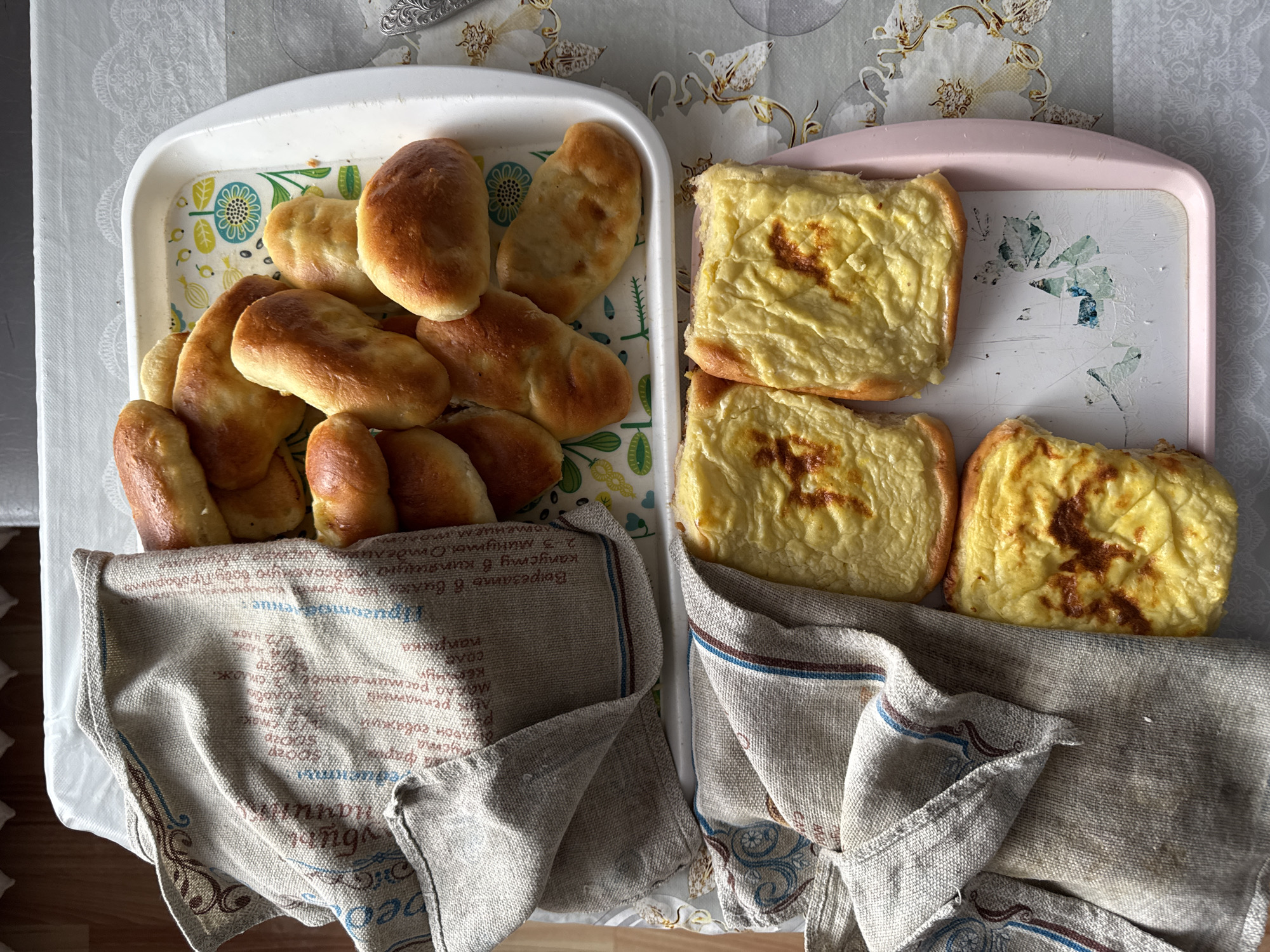 This is a top-down, eye-level shot of two trays of baked goods on a table, likely in a home kitchen. The left tray, which is white, holds a pile of golden-brown baked buns, some of which are covered by a folded linen cloth with a pattern of text and decorative elements. The right tray, which is pink, contains three pieces of a baked casserole or pie with a golden-yellow topping, also partially covered by a similar linen cloth. The background appears to be a tablecloth with a subtle floral pattern. There are no people visible in the image. The image is well-lit and the focus is sharp. It is not possible to determine the exact location within Borodulino, Russia, from the visual information provided.