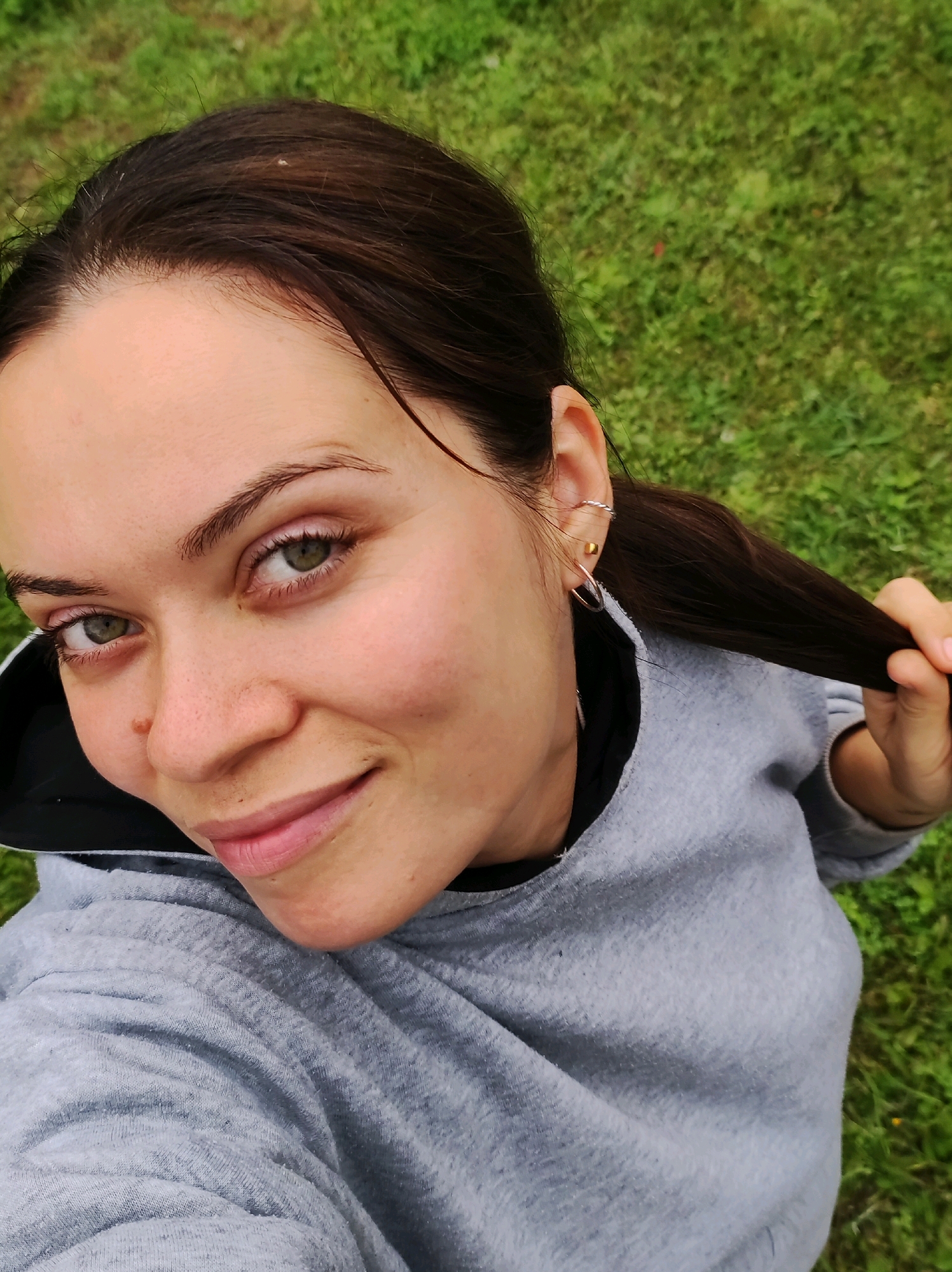 A selfie is taken from a high angle, looking down at a young Caucasian woman with brown hair and green eyes. She is smiling and pulling a strand of her hair. She is wearing a gray hooded sweatshirt. The background is a field of green grass. There are no discernible landmarks to identify the location as Фоки, Russia.