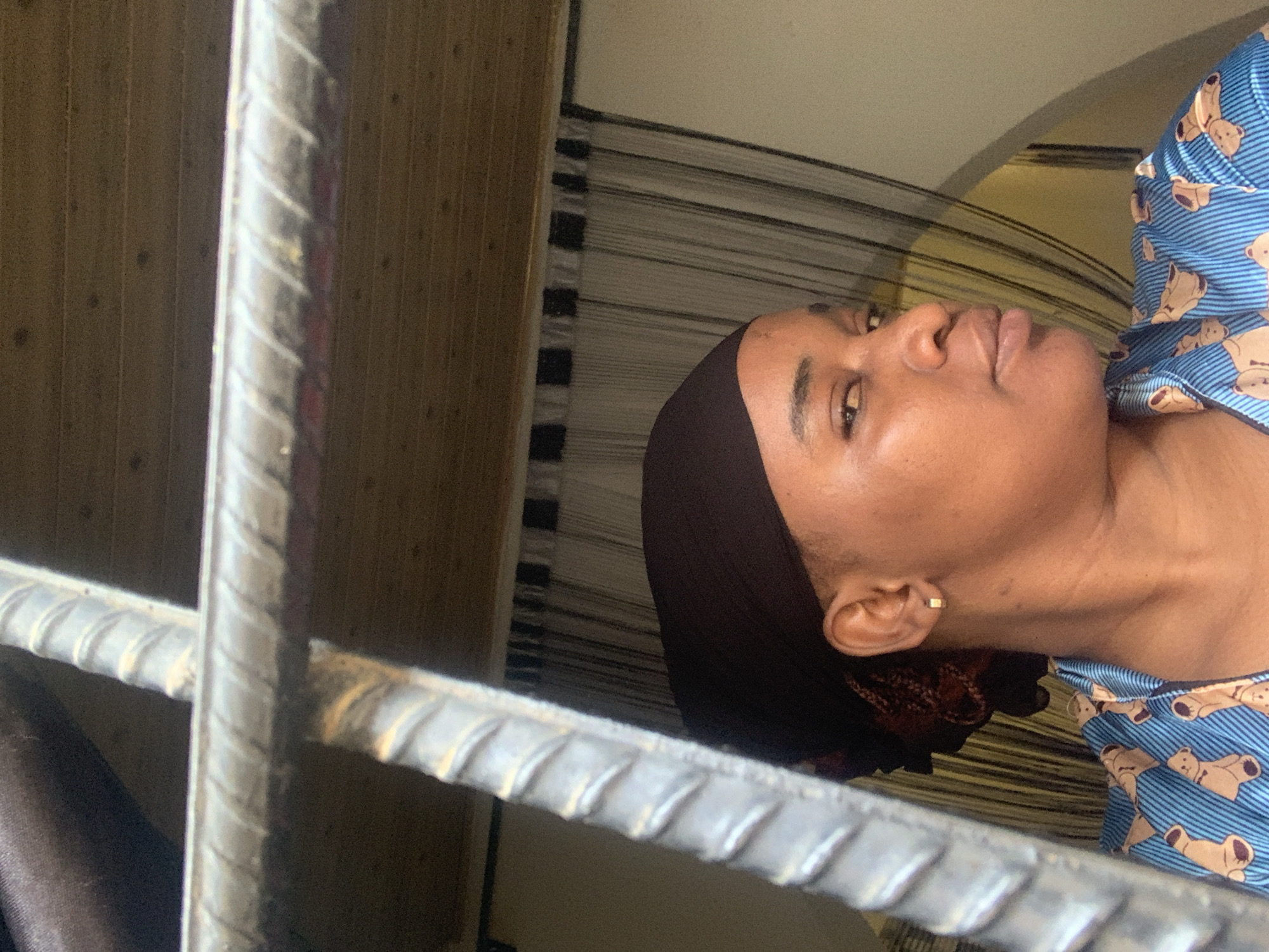 The image captures a profile view of a woman, appearing to look to her right, in an indoor setting within Barakin Akawo, Nigeria. Her dark skin is prominent on her face, neck, and right ear, which features a small, square, gold-colored stud earring. She wears a dark, possibly black or dark brown, head covering. Her attire includes a light blue top patterned with brown teddy bears and thin white horizontal stripes. The woman's expression is neutral and observant.
The foreground is significantly occupied by several metallic, ribbed bars, strongly resembling rebar, which form a partial grid, suggesting security bars on a window or a protective barrier. These bars are light grey with textured surfaces and minor blemishes. Behind the rebar on the left, a light brown, vertically textured surface, possibly wood paneling, is visible. The background directly behind the woman includes a horizontally striped curtain or fabric, with a lighter-colored wall or ceiling above it. The scene is illuminated by soft, natural light, indicating it is daytime. There is no visible text in the image.