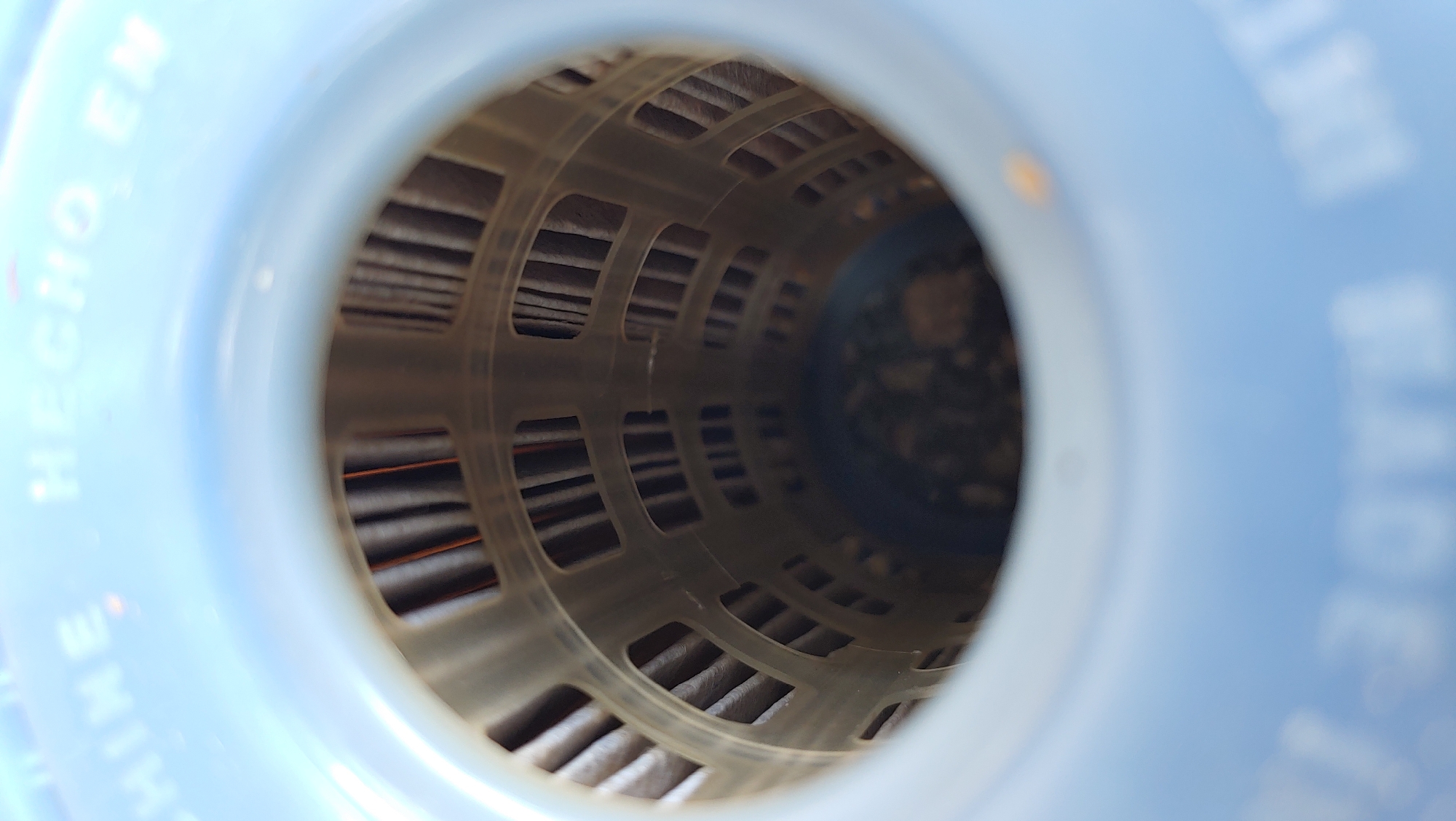 The image is a close-up, interior view of a light blue plastic container. The container has text embossed on its upper rim. Part of the text reads "CHINE HECHO, EN". Inside the container, a cylindrical filter element is visible. This filter is made of a translucent, light-colored plastic cage containing vertical, dark brown filter material. The filter cage has a pattern of rectangular openings. At the bottom of the container, a dark, granular substance is visible, appearing to be dirt or debris. The scene is indoors or outdoors, as suggested by the lighting and the presence of what appears to be natural light on the upper rim. There are no people or animals visible in the image. The location in Mukachevo, Ukraine, cannot be determined from the visual information provided in the image.