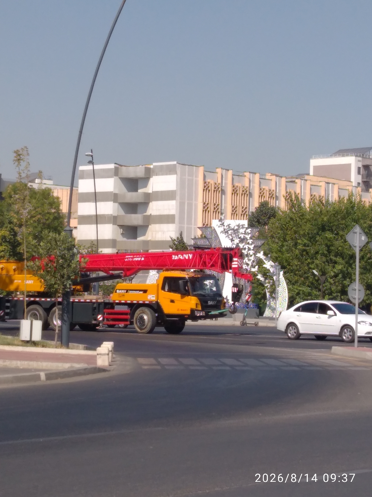 The image depicts a street scene in Tashkent, Uzbekistan, on August 14, 2026, at 09:37. In the foreground, a paved road with a pedestrian crossing is visible. To the left, a yellow and black SANY mobile crane is parked on the street. The crane has a long red boom extended. Behind the crane, trees and modern buildings with light-colored facades and decorative elements are visible. Further to the right, a white sedan is driving on the road. A street light is also present in the scene. The sky is clear and blue.