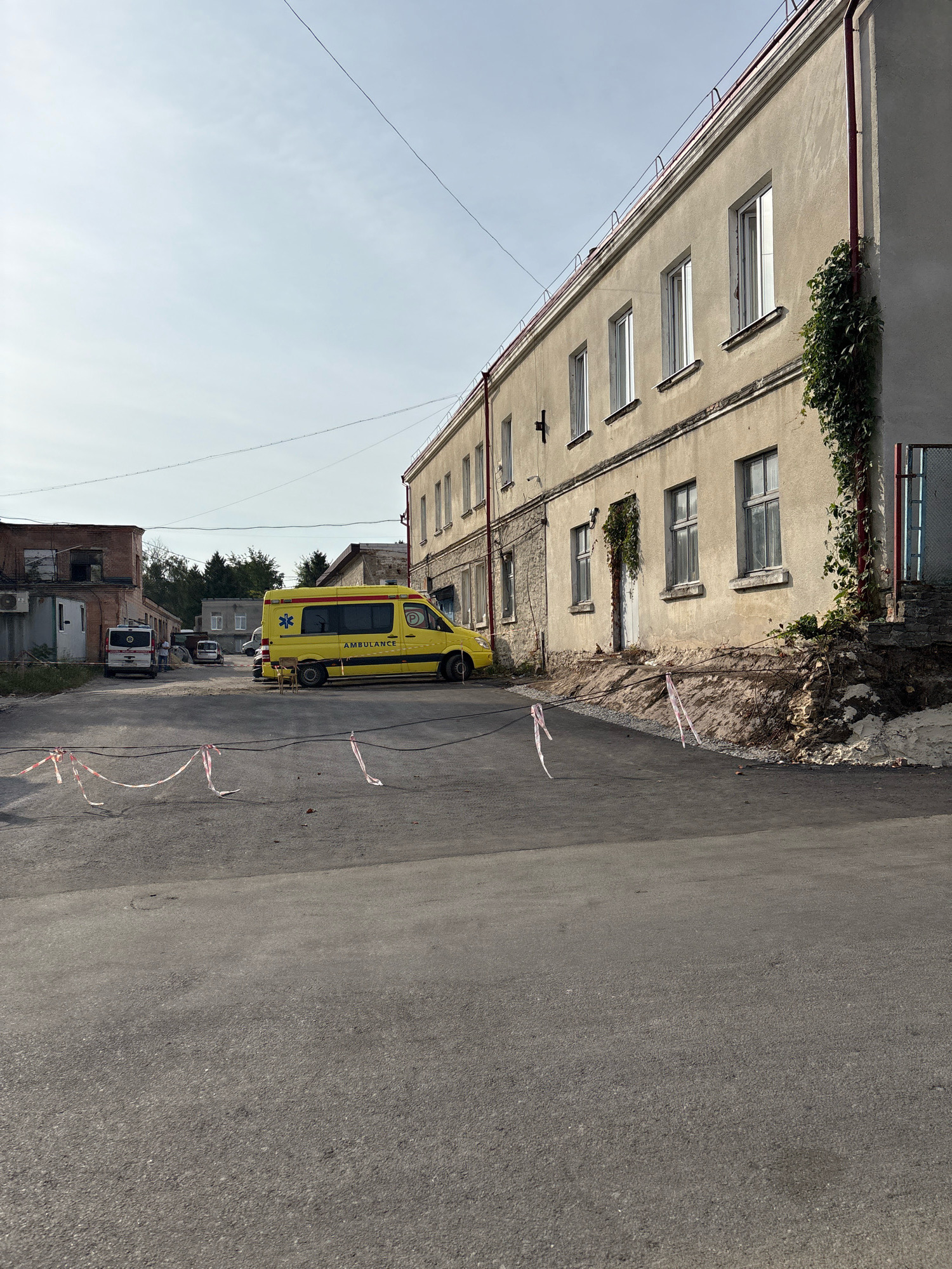 The image depicts an outdoor scene in Ternopil, Ukraine, with a long, two-story building dominating the background. The building has a light-colored facade with numerous windows, some of which are open. Ivy climbs the side of the building in several places. In front of the building, an ambulance van, which is yellow and white, is parked. The van has the word "AMBULANCE" written on its side. A white van is parked further to the left. The ground in front of the building is paved with asphalt. Red and white caution tape is strung across the foreground, indicating a restricted area. The sky is overcast, and the overall impression is one of an ordinary day in an urban environment.