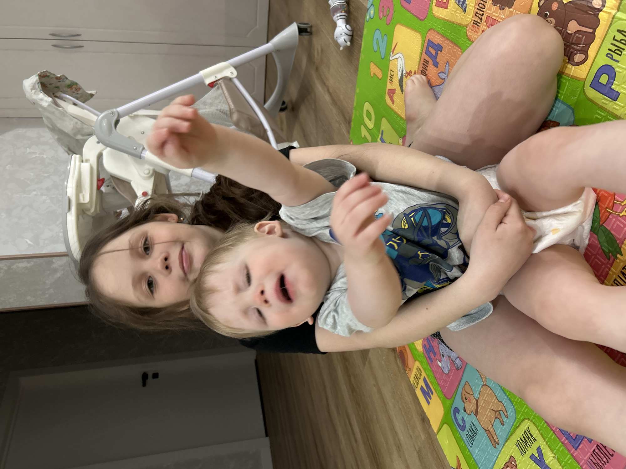 The image depicts two children, a girl and a baby, sitting on a colorful play mat in a room in Chaikovsky, Russia. The girl, presumably older, is holding the baby, who appears to be fussing or crying, in her lap. The girl is looking at the camera with a slight smile. The play mat is patterned with Cyrillic letters and animal images. A baby walker is partially visible in the background, and there is a white door or cabinet in the background as well.