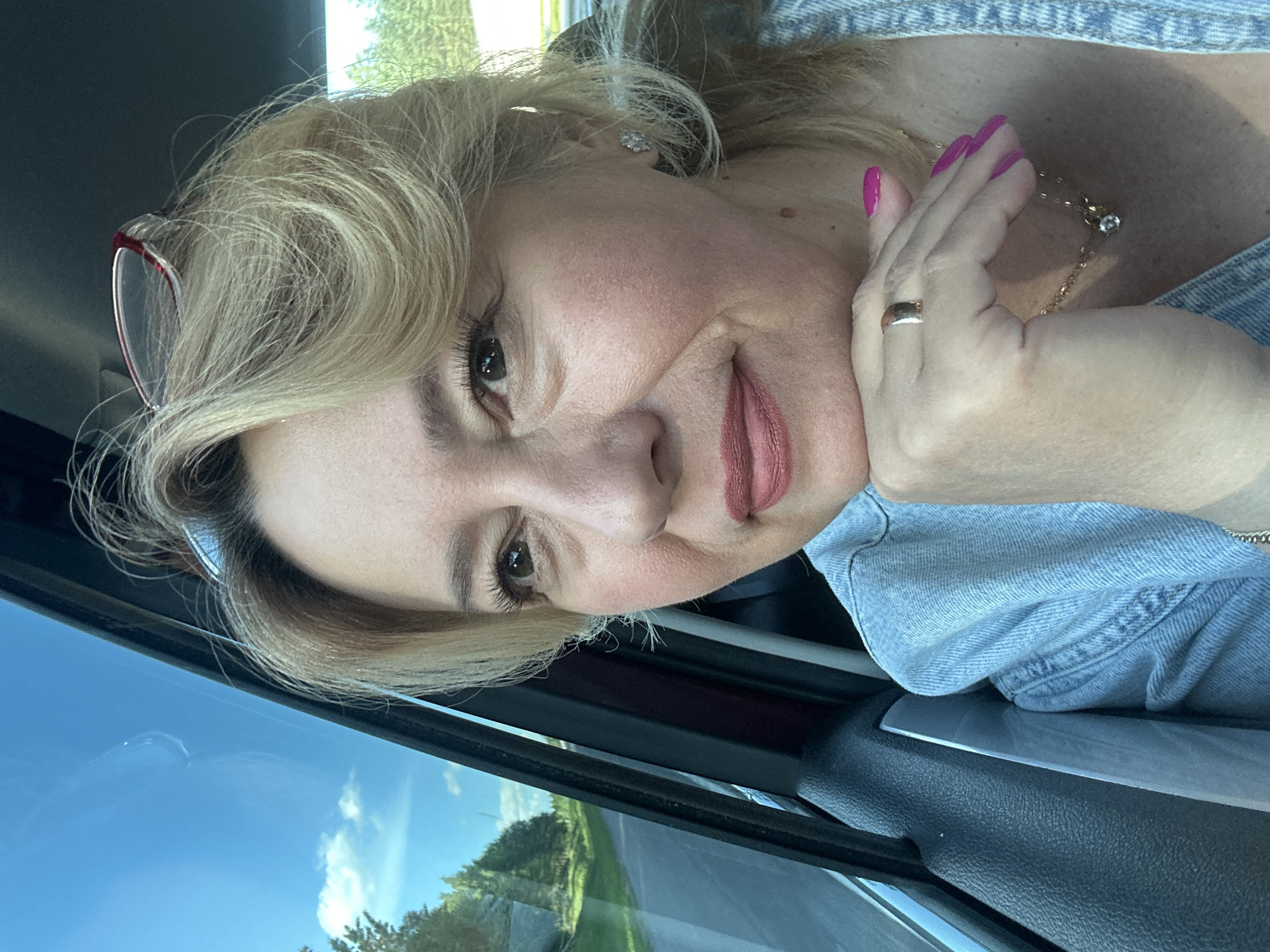 A blonde woman is in the passenger seat of a car, looking out the window. She is wearing a light blue denim jacket and has pink nail polish. Her hand is resting on her chin, and she is wearing a ring and a necklace. The window shows trees and a road, suggesting the car is on a rural road. The sky is blue with some clouds. The image is a selfie taken from a slightly high angle.