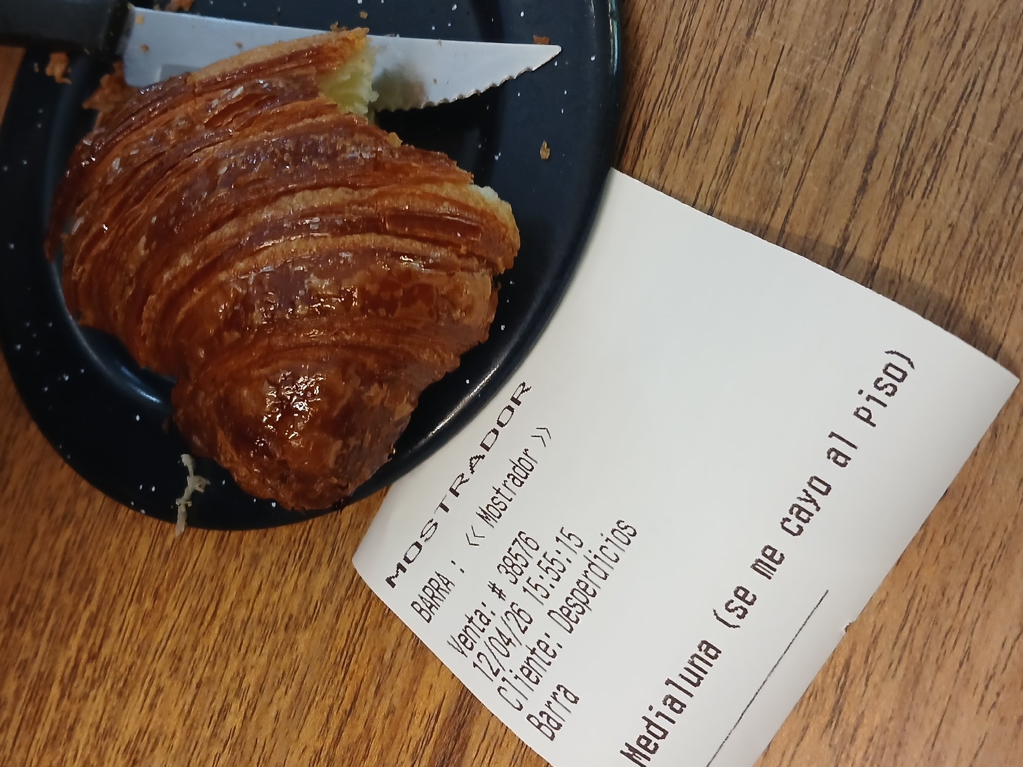 On a wooden surface, a partially eaten croissant rests on a small, black speckled plate next to a printed receipt. The receipt, angled slightly towards the viewer, lists items including "Medialuna (se me cayo al piso)" (croissant (I dropped it on the floor)), indicating a write-off due to it being dropped. Further text on the receipt indicates "MOSTRADOR" (counter), "BARRA : <<< Mostrador >>" (bar), "Venta: # 38576" (sale), "12/04/26 15:55:15", "Cliente: Desperdicios" (customer: waste), and "Barra." A knife with a white serrated blade rests on the plate with the croissant. The scene suggests a cafe or eatery in Buenos Aires, Argentina.