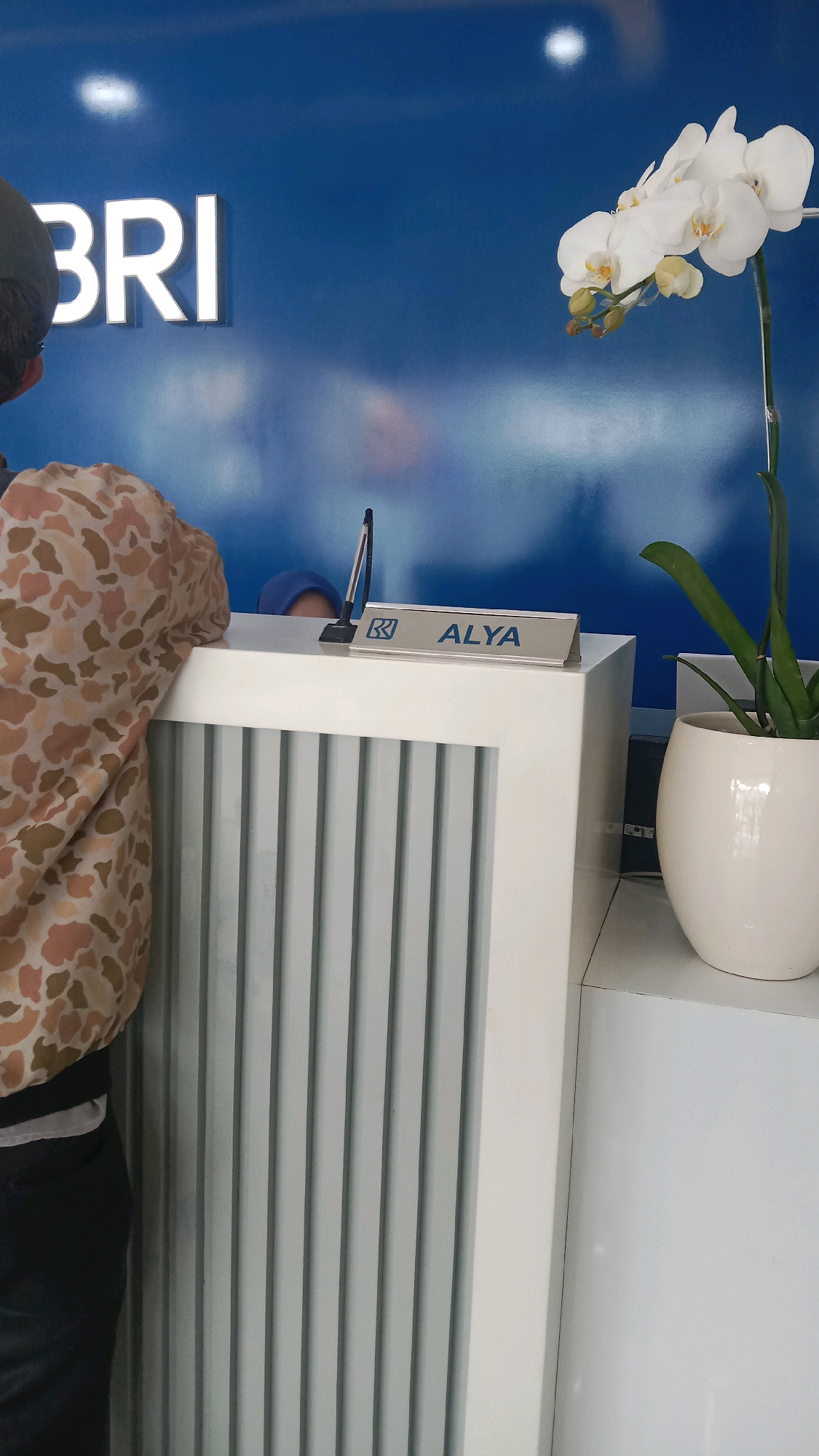The image depicts the interior of a BRI bank branch in Bekasi, Indonesia. In the foreground, a person wearing a camouflage-patterned shirt and dark pants stands at a counter, facing away from the camera. Behind the counter, a bank teller wearing a blue hijab is partially visible. A small sign on the counter reads "BRI ALYA." A white orchid plant in a pot sits to the right of the counter. The bank's blue logo "BRI" is prominently displayed on the wall behind the counter. The environment appears to be a typical bank reception area with a clean, modern aesthetic.