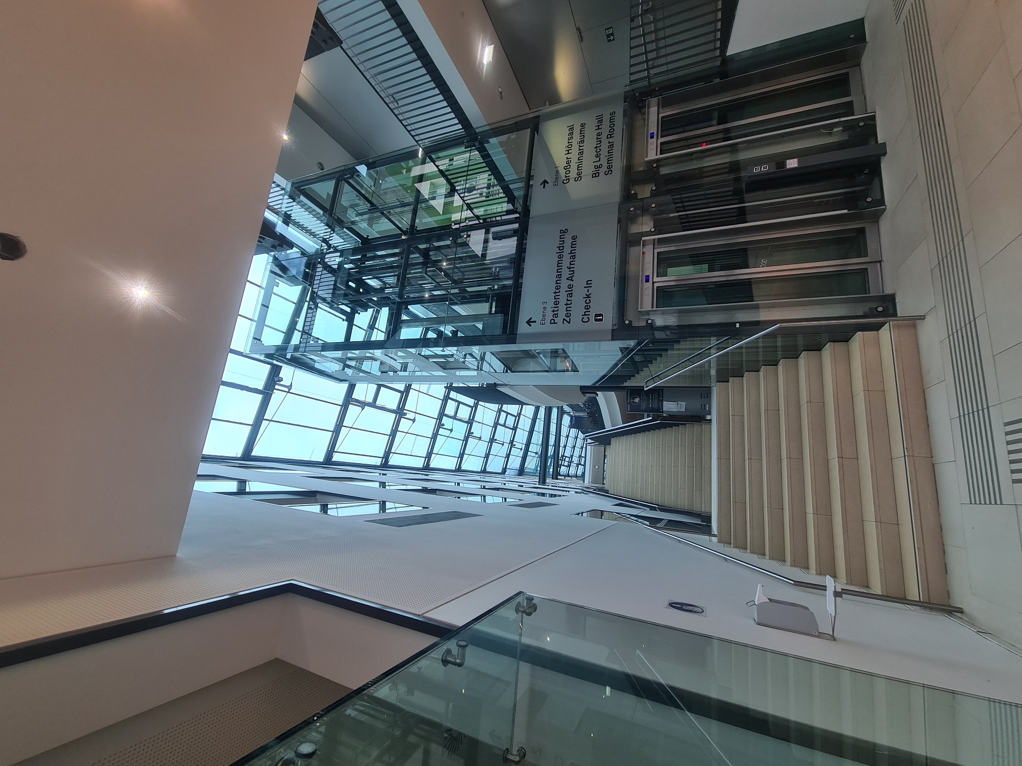 The image shows the interior of a modern building, likely a hospital or medical facility, featuring a central atrium with a glass ceiling and a glass-enclosed elevator. A wide staircase made of light-colored stone ascends on the left side of the frame, with a glass railing. The walls are painted white, and there are directional signs in German and English indicating "Ebene 3 Patientenanmeldung Zentrale Aufnahme Check-in" (Level 3 Patient Registration Central Admission Check-in) and "Ebene 1 Großer Hörsaal Seminarräume Big Lecture Hall Seminar Rooms". The elevators are also made of glass and steel, with digital displays showing floor numbers. The overall impression is one of spaciousness and natural light, with a clean and functional design. While the city of Vienna is mentioned, the image itself does not contain any specific landmarks or features that would pinpoint its exact location within the city.