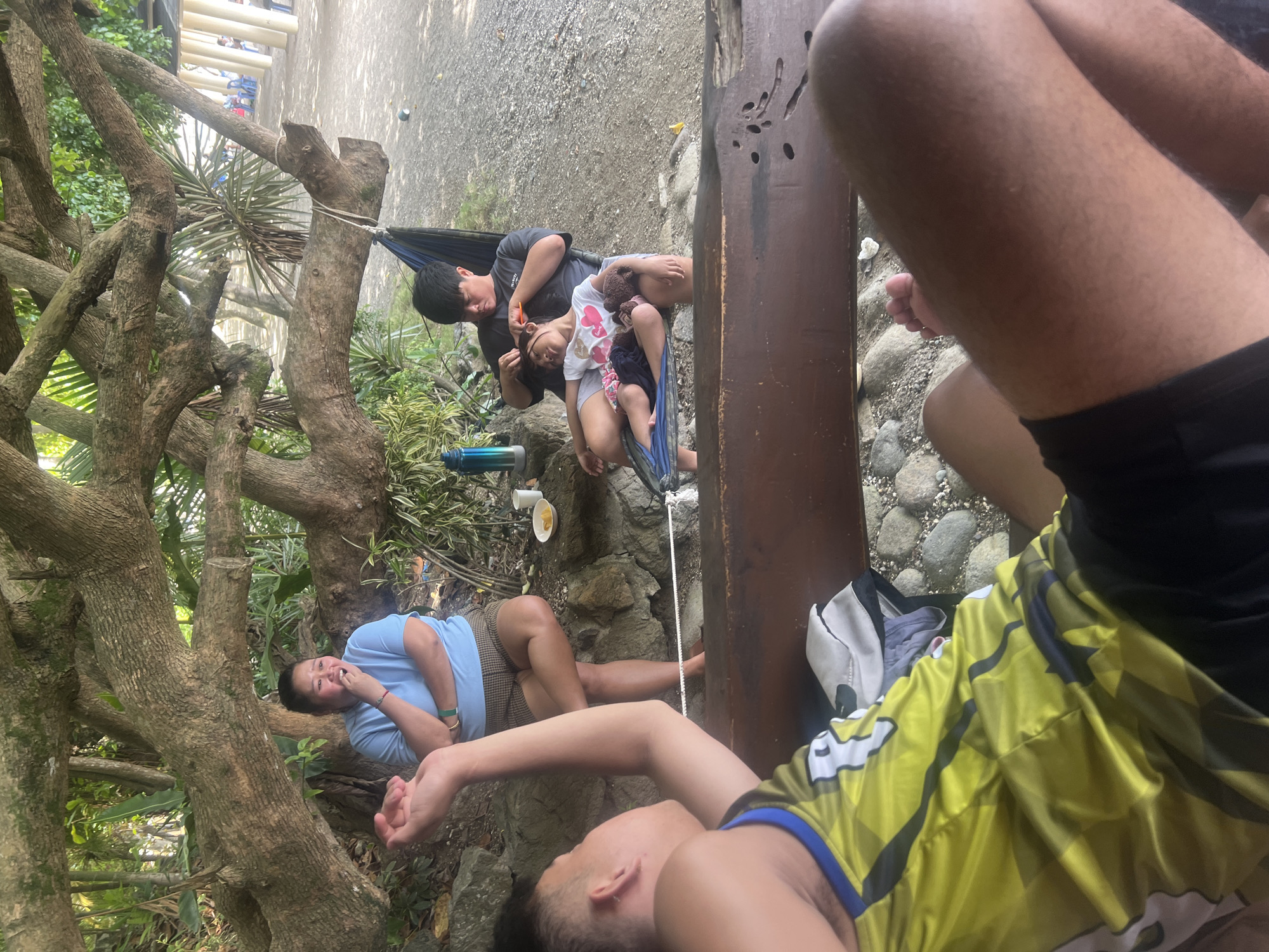 The image shows a group of people relaxing outdoors in Banaybanay, Philippines. A hammock is strung between a tree and a wooden post. A man is in the hammock with a child sitting on his lap. Another person is lying on the ground near the hammock. A second person is sitting next to the post. There is another person in the lower part of the frame. The ground is rocky. A white bowl is next to the hammock.