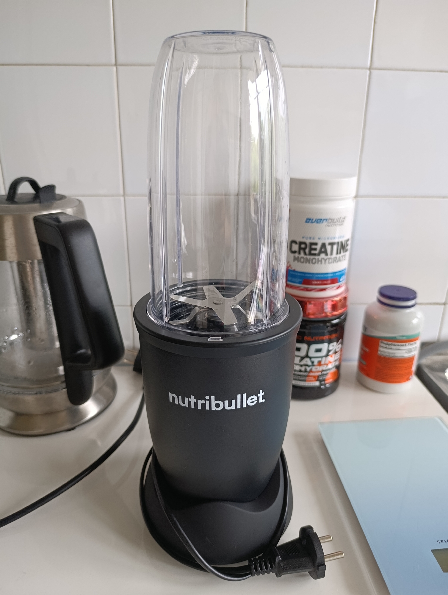 An overhead, medium shot captures a NutriBullet blender on a white countertop, positioned in front of a tiled wall. To the left of the blender, a silver electric kettle with a black handle is visible. Behind the blender, a stack of three supplement containers are arranged, with the top one labeled "EVERBUILD CREATINE MONOHYDRATE." To the right of the supplements, a white bottle with an orange label, likely a supplement, stands. A portion of a light blue digital scale is visible in the lower right corner of the frame. The blender's power cord and plug are lying on the countertop. The scene appears to be in a kitchen or pantry setting.