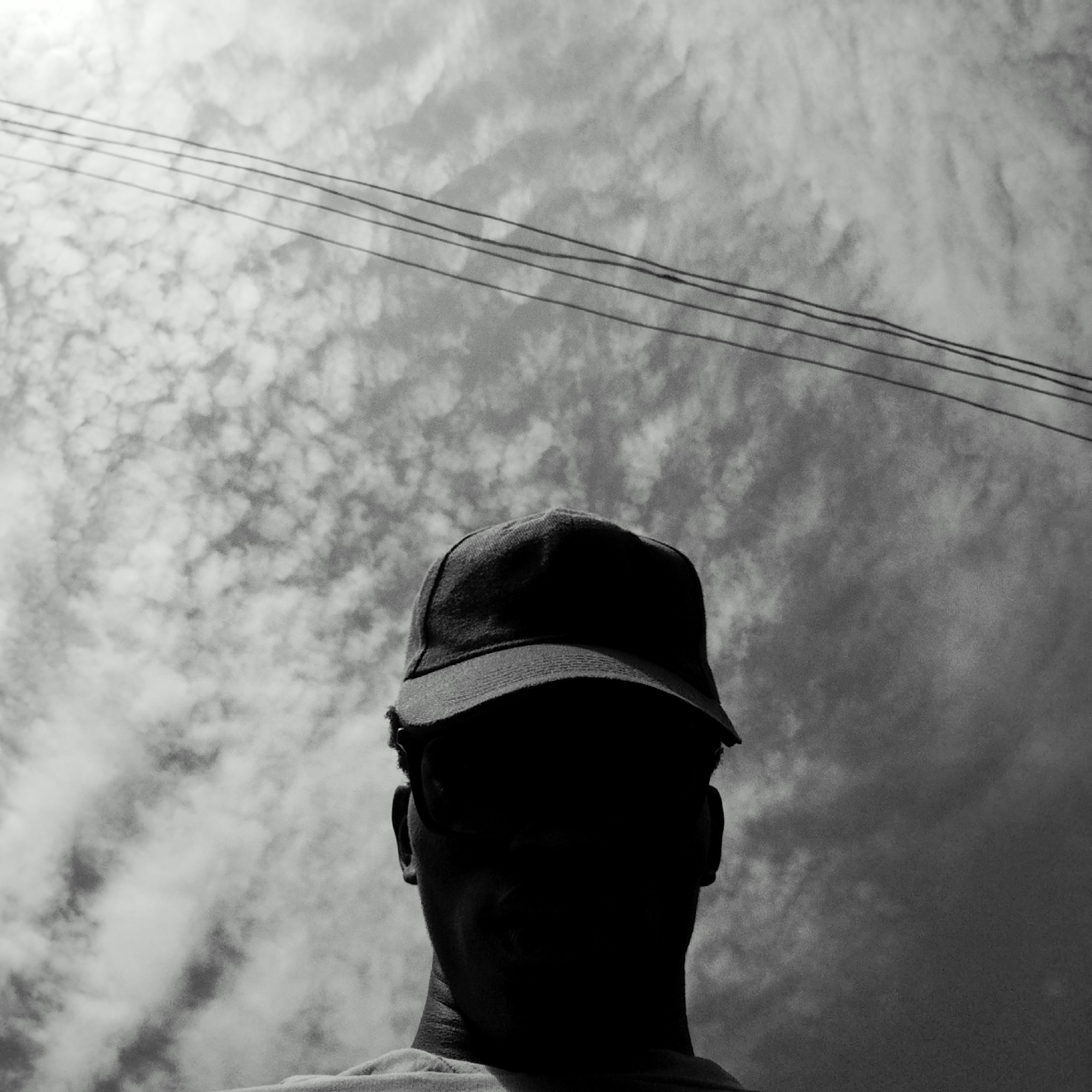 A black and white, low-angle shot shows a person wearing a baseball cap with their face in shadow. Overhead are multiple power lines against a cloudy sky with a textured cloud formation. The location is unknown.