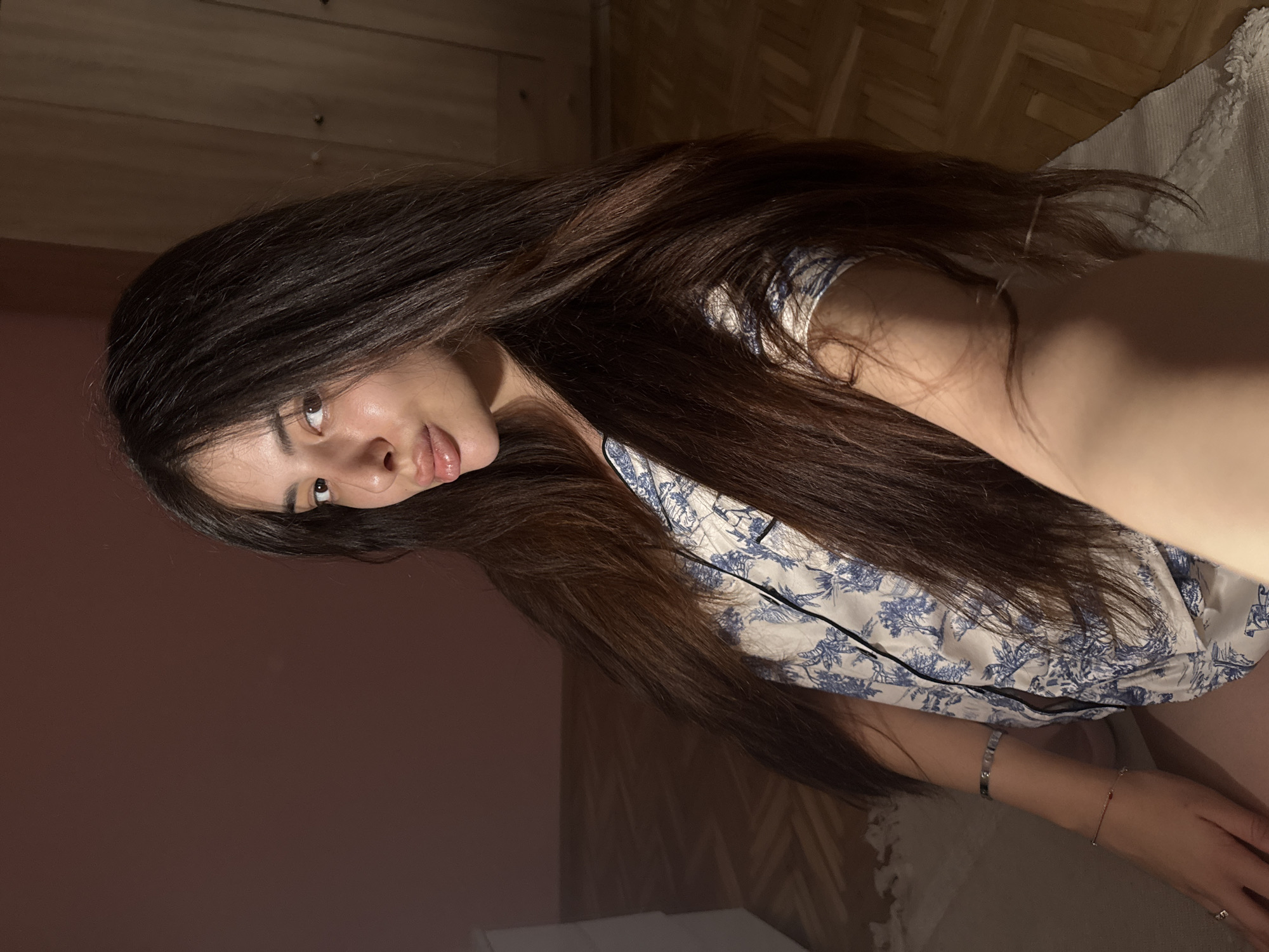 A young woman with long, dark brown hair is seated indoors, facing the camera. She is wearing a blue and white patterned top. Her lips are slightly parted, and she is looking directly at the camera. Her left arm is visible, resting on her lap, and she is wearing a bracelet. The background consists of a wooden floor and a wall that appears to be painted pink. A wardrobe is visible on the right side of the image. The lighting suggests an indoor setting, possibly a bedroom or living area. No specific location in Plovdiv, Bulgaria can be identified from the image.
