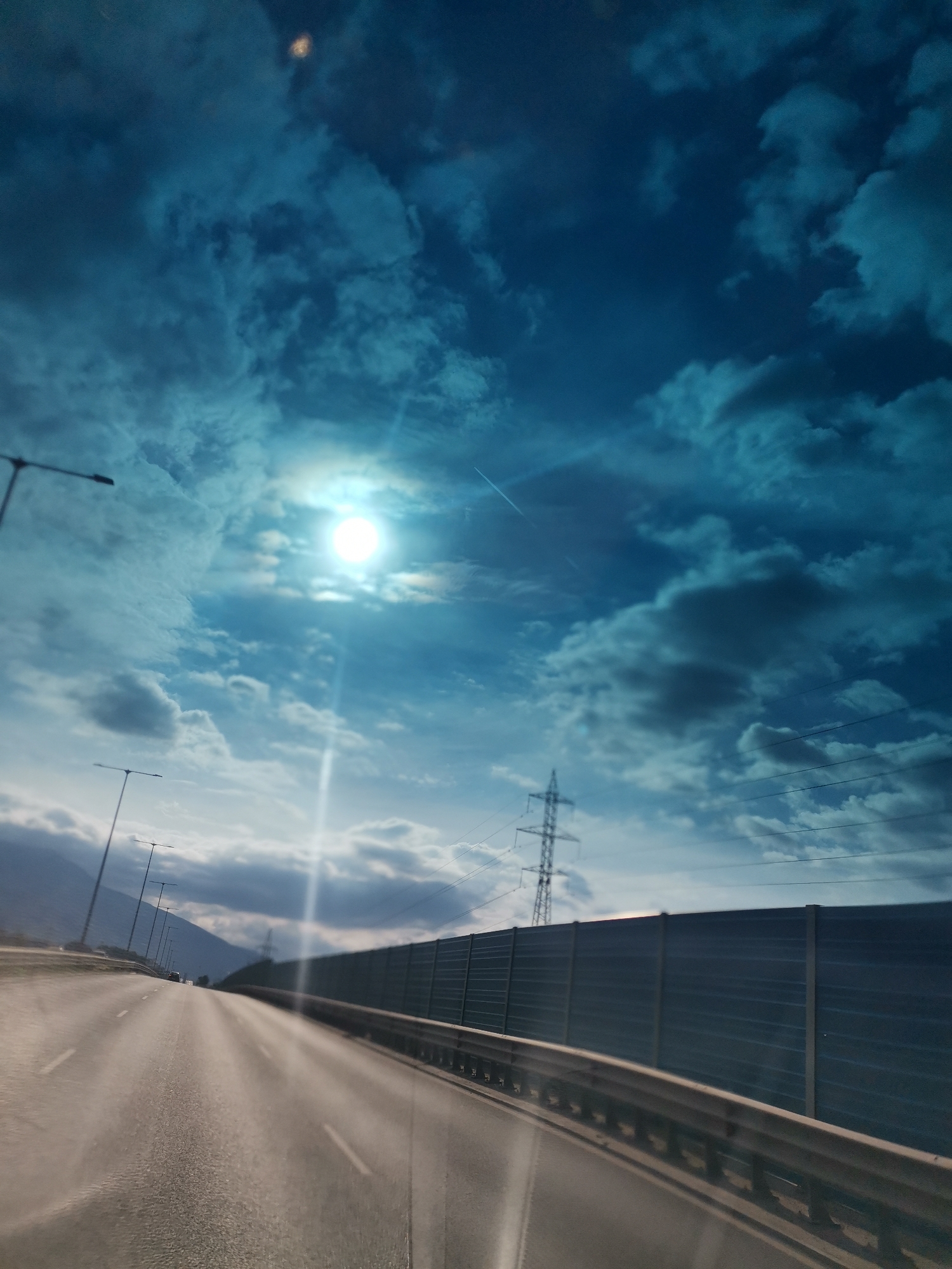 The image shows a view from a vehicle traveling on a highway. The highway is paved and has lane markings. On the right side of the highway, there is a sound barrier. In the distance, there are mountains. The sky is cloudy with the sun visible, creating lens flare. There are also streetlights and a power line tower visible. The overall scene appears to be an urban or suburban area with infrastructure for transportation and electricity. There are no people or animals visible in the image.