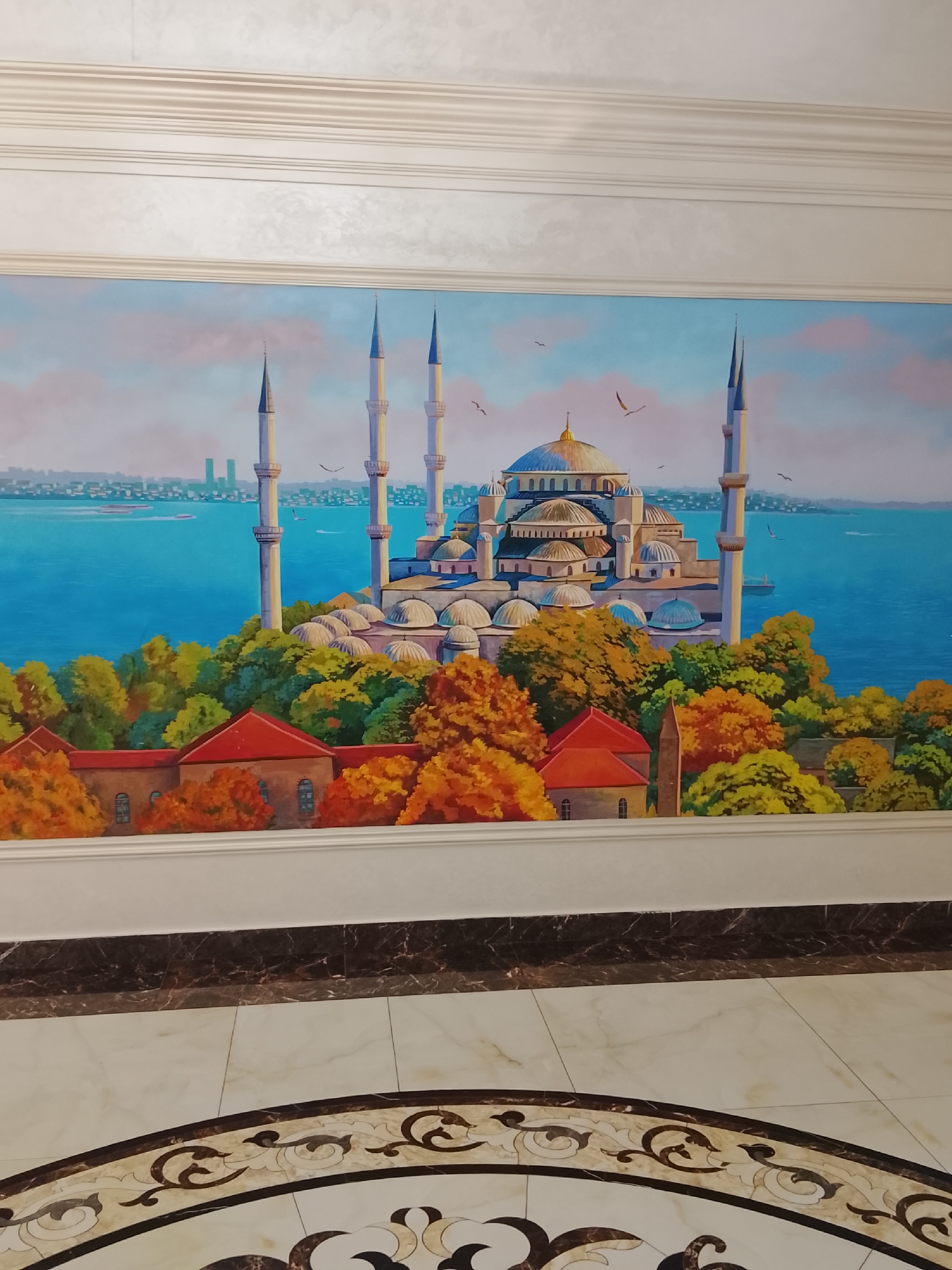 A mural depicts the Blue Mosque in Istanbul, Turkey, set against a backdrop of a bright blue sea and sky with scattered clouds and birds. The mosque itself is richly detailed with a large central dome, multiple smaller domes, and tall, slender minarets. In the foreground, lush trees with vibrant green and orange foliage surround a series of buildings with red tile roofs. The mural is bordered by white molding at the top and a white baseboard at the bottom. Below the mural, a marble floor with a decorative inlay pattern is visible, featuring a combination of light beige tiles and darker, intricate designs. The scene is static and appears to be an interior decoration.