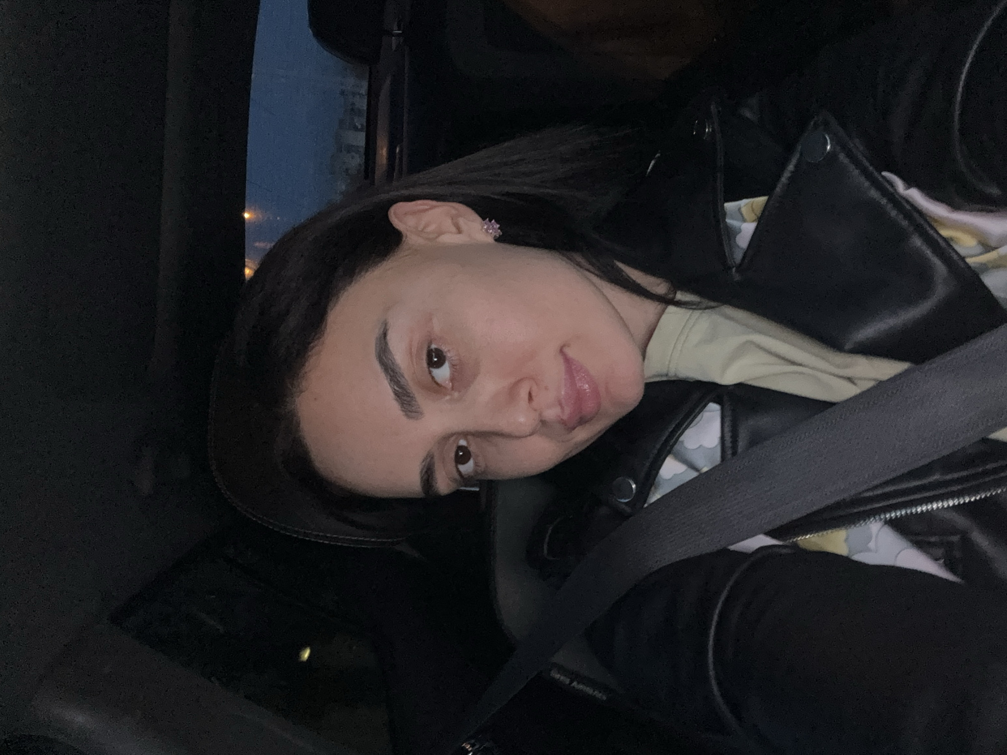 A casual self-portrait captures a young woman inside a car in Tbilisi, Georgia, during the evening or night. She has long, dark hair, dark, well-defined eyebrows, and brown eyes, looking directly at the camera with a subtle smirk or pout. She is wearing a black leather-look jacket over a light-colored top, which appears to have a soft, muted pattern underneath the jacket. A small, star-shaped earring, possibly pink or purple, is visible in her right ear.
She is seated in the car, with a black seatbelt fastened across her chest. Partial text, "SRS Airbag," is visible on the seatbelt strap or buckle area. The car's interior is dimly lit, primarily by ambient light. Through the window in the background, a dark blue sky with scattered distant artificial lights suggests an urban environment after sunset. The overall impression is informal and relaxed.