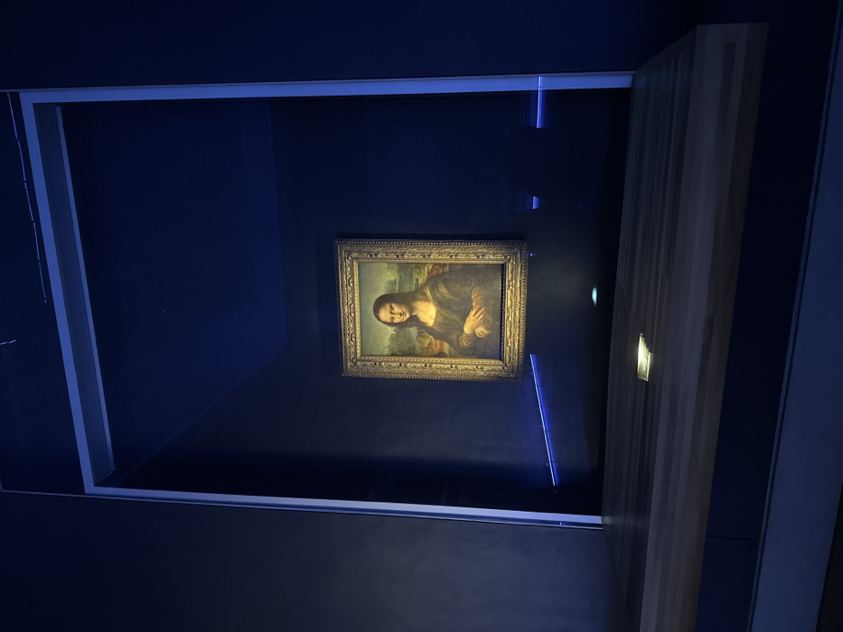 The image displays the painting "Mona Lisa" (La Gioconda) by Leonardo da Vinci, centrally located within a dimly lit exhibit space at the Louvre Museum in Paris, France. The painting is encased behind a protective glass barrier and framed by an ornate gold-toned frame. It is individually illuminated by direct spotlights, contrasting with the dark blue walls of the room. A pervasive blue ambient light emanates from linear sources, casting a blue hue throughout the environment. In the lower foreground, a light-colored wooden barrier or display counter is present, featuring a small, rectangular reflective element on its surface. The painting is situated within a recessed opening defined by light-colored vertical and horizontal structural elements. No individuals or actions are visible within the scene.