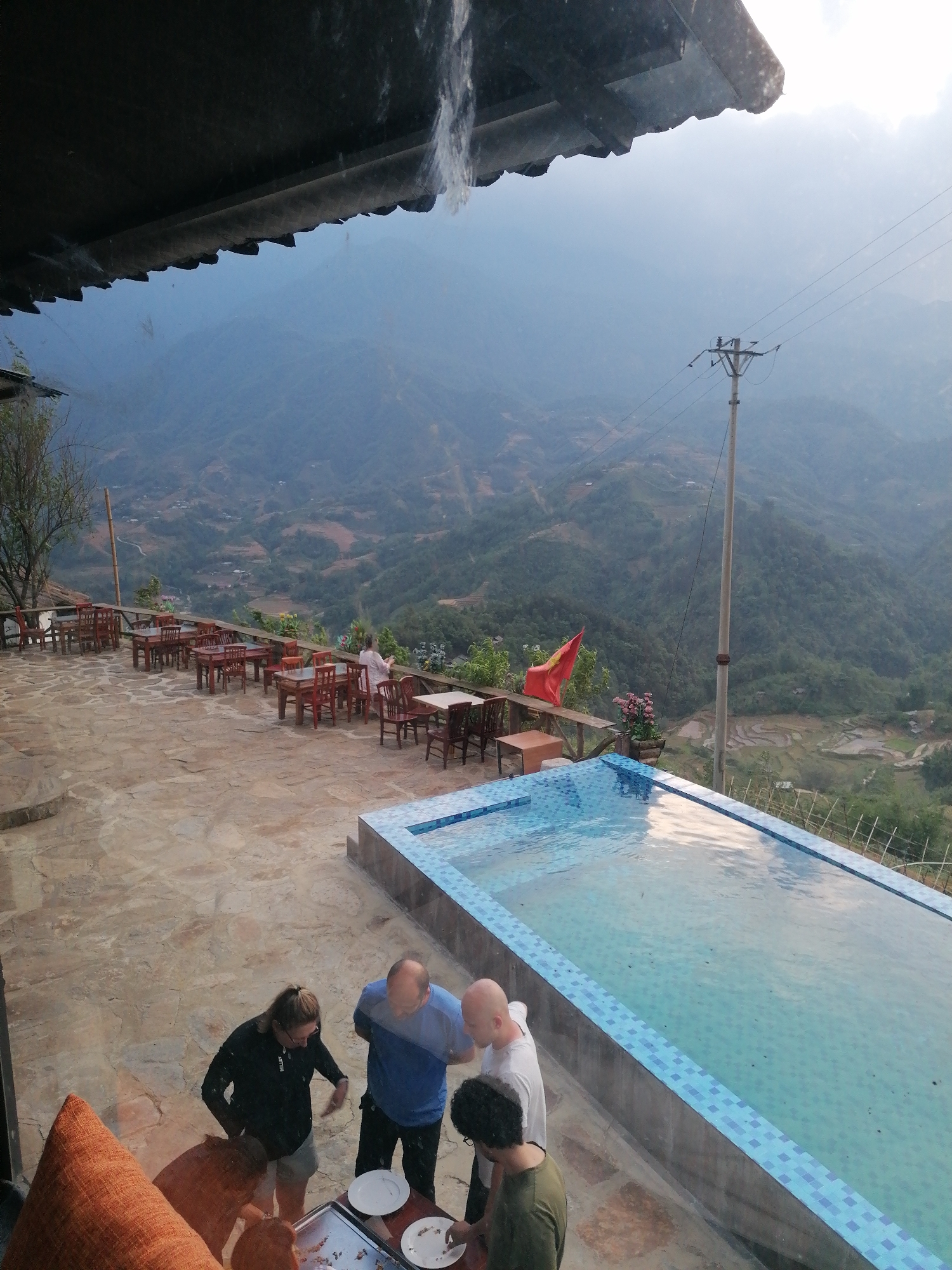 The image captures a panoramic view from an elevated outdoor terrace, likely part of a resort or restaurant, overlooking the vast, hazy mountainous landscape of Sa Pa, Vietnam.
In the immediate foreground, part of a dark overhang casts a shadow, and an orange cushioned seat is visible on the left. On the stone-paved patio, four individuals are gathered around a table, seemingly serving themselves from dishes, possibly during a meal or buffet. A woman in a dark top and shorts stands looking at the table, while a man in a blue shirt and another bald man in a white shirt stand nearby. Another person with dark curly hair is partially visible on the right.
The patio extends further, featuring an outdoor dining area furnished with numerous wooden tables and matching chairs arranged in rows, all facing the scenic valley. A person in white is seated at one of these tables in the mid-ground, enjoying the view. To the right, a long, rectangular swimming pool with blue and white checkered tiling lines its edges, reflecting the cloudy sky. Beyond the pool and patio railing, a red Vietnamese flag flies from a pole, adding a distinct national touch.
The background is dominated by layer upon layer of lush, verdant mountains, characteristic of Sa Pa's terraced terrain, which recede into a soft, atmospheric haze. Valleys below reveal patches of rice paddies and scattered structures, indicating rural settlements. A tall utility pole with power lines is prominent on the right, extending into the distant landscape. The overall lighting suggests either a soft morning light or late afternoon, with an overcast or hazy sky creating a diffused illumination across the scene. There is no visible text.