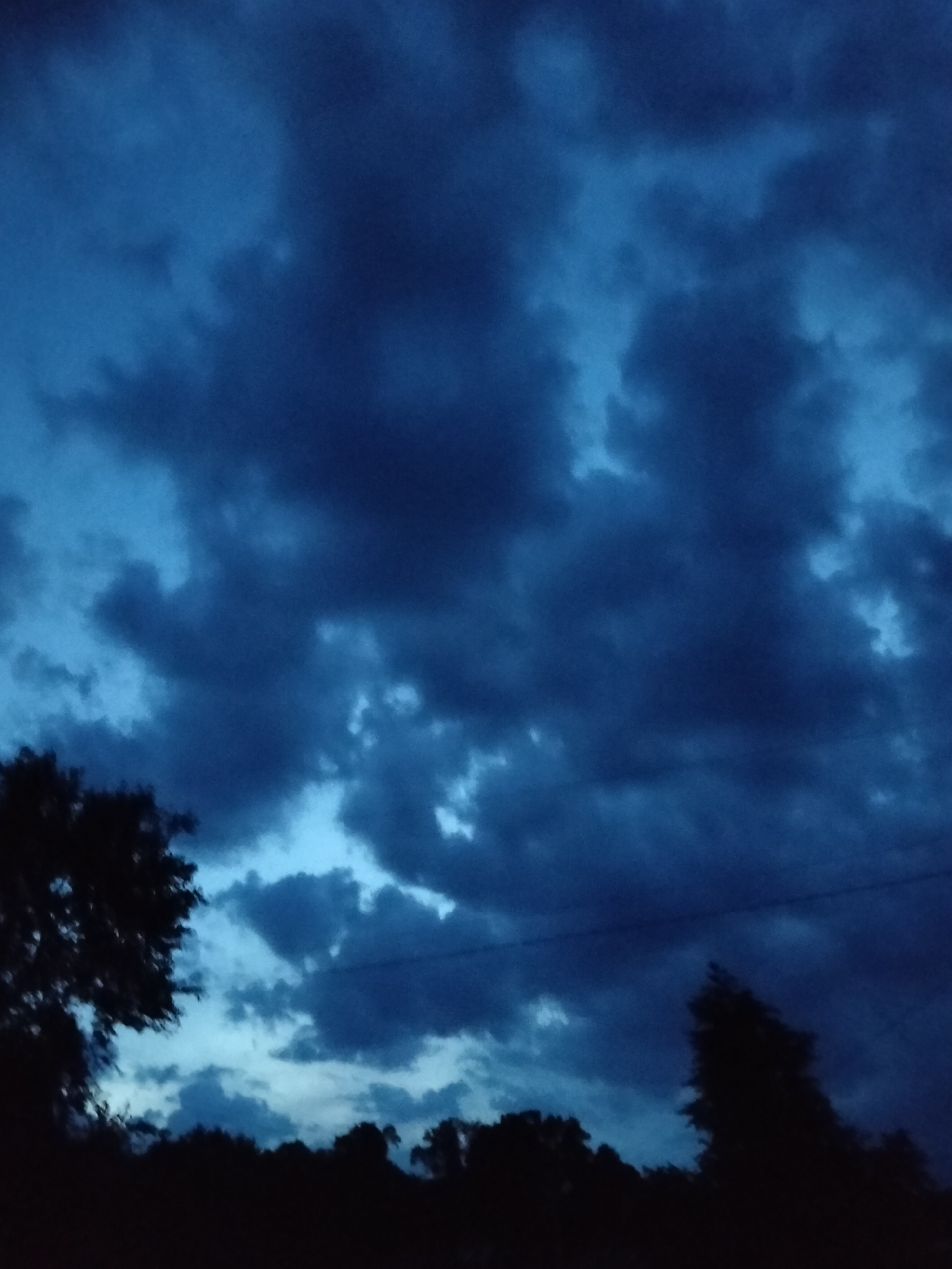 This is a photograph of a dark, cloudy sky over a silhouetted treeline. The sky is a deep, moody blue with darker storm clouds obscuring most of the light. There are lighter patches of sky visible through the clouds, suggesting it is either dawn or dusk. The treeline at the bottom of the image is completely dark, making it impossible to discern any details of the trees or the ground beneath them. The overall impression is one of an approaching storm or the aftermath of one. There is no text visible in the image.