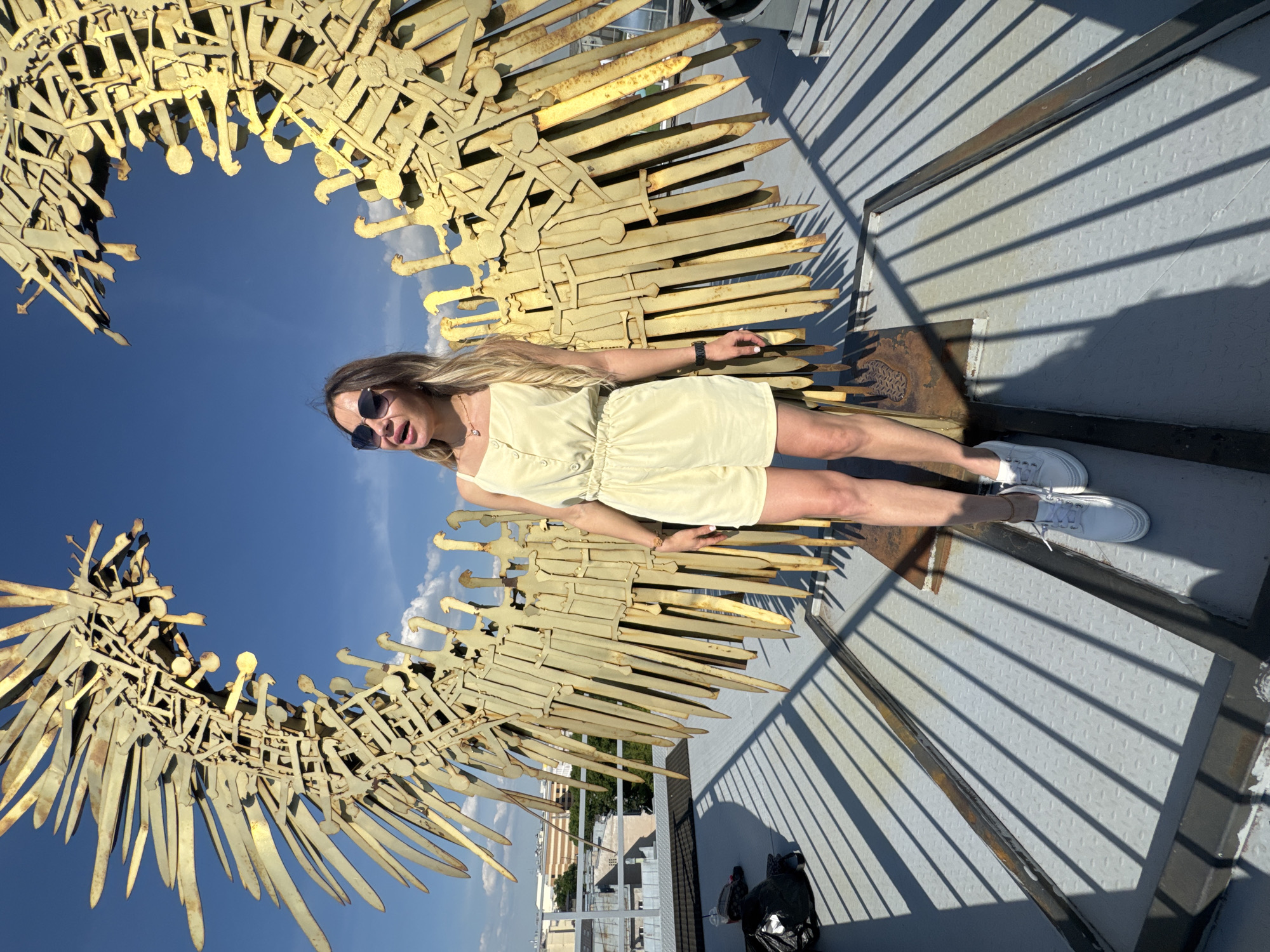 A blonde woman in a yellow romper and white sneakers stands in front of a large, golden, wing-like sculpture made of metal. The sculpture appears to be constructed from many individual pieces, some resembling swords or blades, that are welded together to form a circular shape with jagged edges. The woman is wearing sunglasses and a necklace. The sculpture is situated on a rooftop platform with a grey metal grated floor. In the background, the blue sky with scattered clouds is visible, as are some buildings and trees. The overall setting suggests an outdoor urban environment. This sculpture is located on the rooftop of the Russian Museum in Saint Petersburg, Russia.