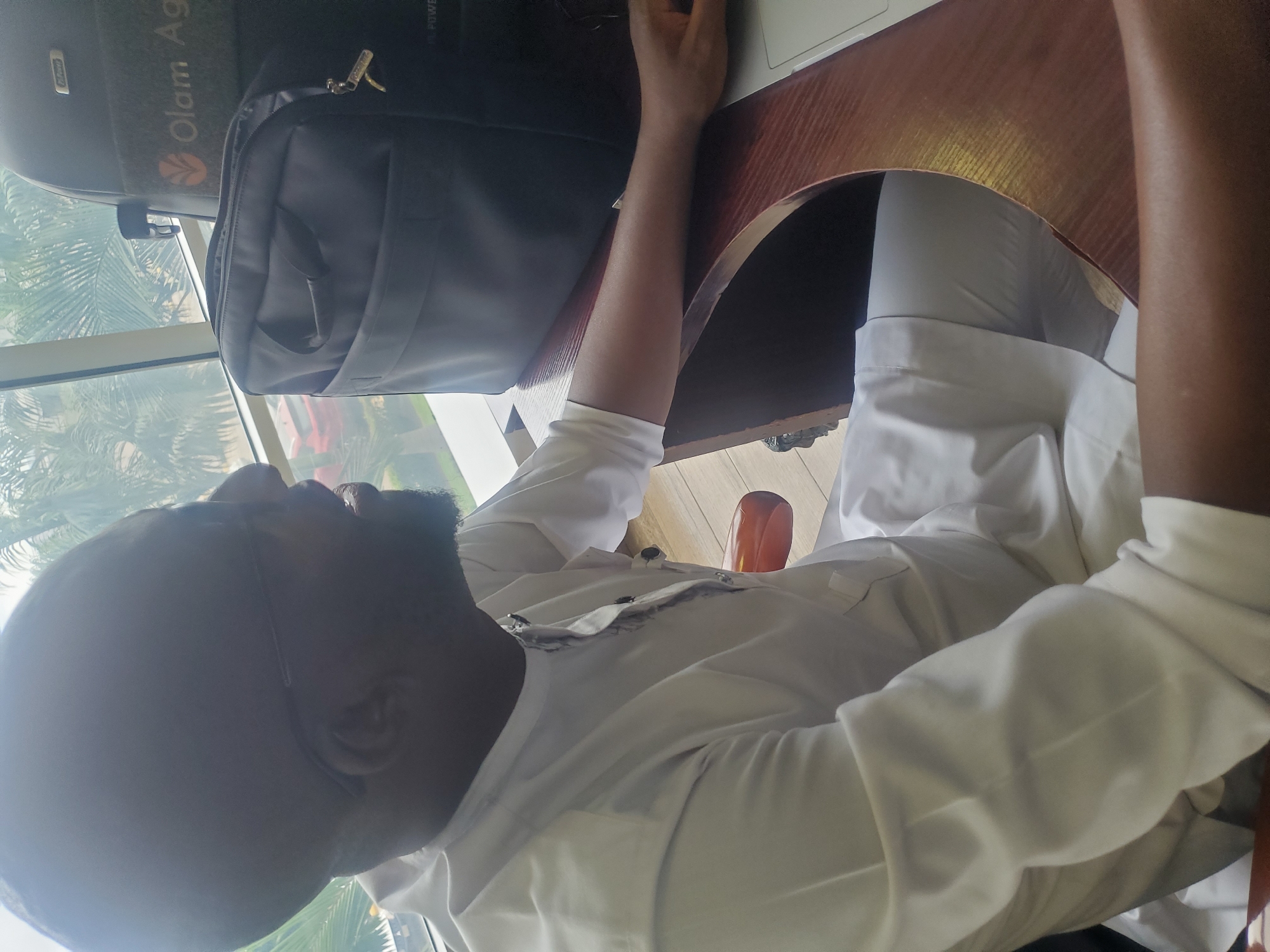 The image captures a person sitting at a wooden desk indoors, looking out a large window during the daytime. The individual has dark skin, a short beard, and appears to be wearing glasses. They are dressed in a light-colored, possibly white, long-sleeved shirt and matching trousers. Their right arm rests on the desk, while their left hand is positioned near an open laptop.
Through the window, bright natural light indicates it is daytime. The outdoor scene features lush greenery, notably palm trees, and a glimpse of a red vehicle in the distance. This tropical or subtropical vegetation aligns with the provided context of Gwagwada, Nigeria.
Adjacent to the person, a black laptop bag or briefcase is placed on top of a larger dark object, which could be a piece of luggage or the back of a seat. A small golden-colored lock or zipper pull is visible on the bag. Clearly discernible white text on the larger dark object reads "Olam Ag", likely referring to Olam Agriculture. Additionally, faint white text resembling "OL. POWER" is visible on the top right edge of the same object. The person's posture appears relaxed, suggesting they are either working, studying, or simply taking a break and observing the view outside. Part of a reddish-brown wooden chair is visible under the desk.